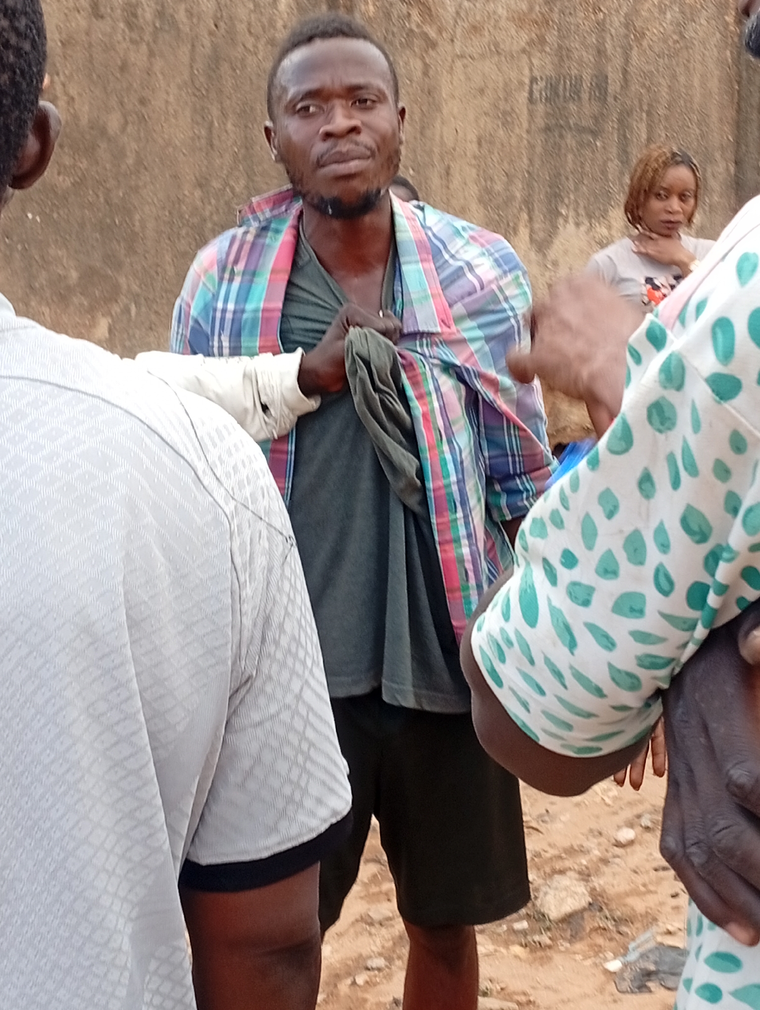 The image captures a scene in Sabon Tasha, Nigeria, depicting a man with a short haircut and beard standing outdoors. He is wearing a gray shirt, black shorts, and a multicolored plaid shirt draped over his shoulders. He appears to be interacting with two individuals who are partially visible on either side of him, obscuring the view. A woman with blonde hair is visible in the background. The setting appears to be an unpaved area with a wall visible behind the man.
