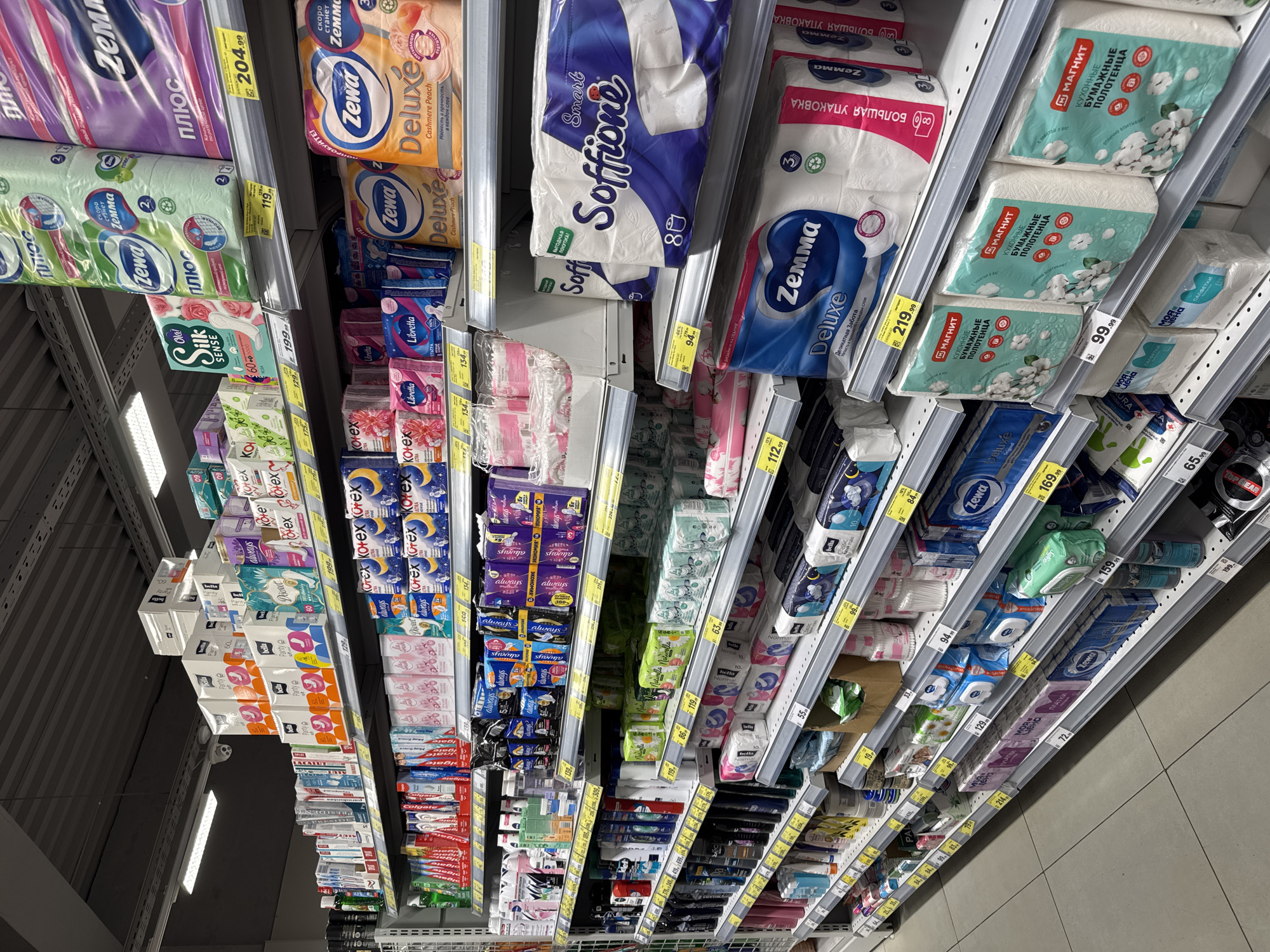 The image depicts a supermarket shelf stocked with a variety of household goods, primarily paper products such as toilet paper, paper towels, and feminine hygiene products. The products are arranged on tiered shelving and bear brand names like Zewa, Soffione, Libretta, Kotex, and Магнит. Price tags are visible below each product, indicating the cost in Russian rubles.
The brands suggest the location is in Russia. The language on the product packaging and price tags confirms this is Saint Petersburg, Russia.