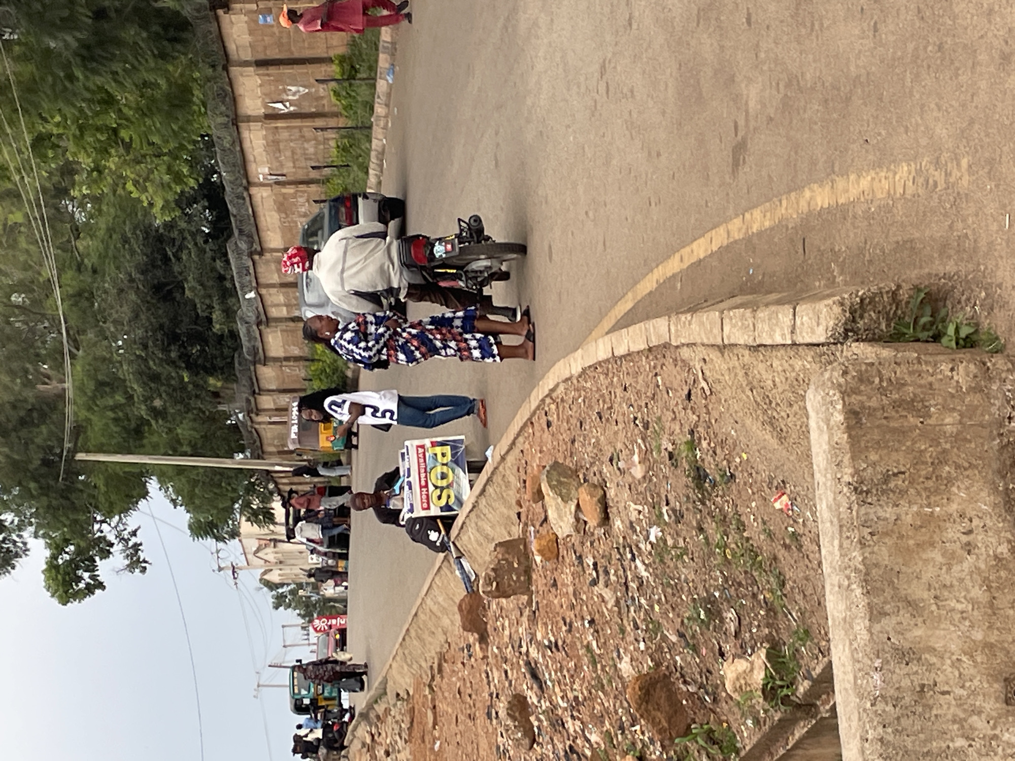 The image depicts a street scene in Jos, Nigeria, with a focus on individuals and their activities. In the foreground, a dirt roadside is visible, with scattered rocks and debris. A concrete structure runs along the edge of the road.
Further down the street, a man is seated behind a stand displaying a "POS Available Here" sign, indicating a point-of-sale service. Beside him, a woman in a patterned dress stands. A man on a motorcycle is positioned behind them, facing away from the camera, suggesting he might be a passenger or waiting.
In the background, several other people are present, some standing and others walking. Various vehicles, including a tricycle, cars, and a motorcycle, are seen on the road. Buildings and trees line the street, and a high wall with barbed wire tops it, suggesting a secure area or property. The sky is overcast, contributing to the generally muted lighting of the scene. The overall atmosphere appears to be that of a bustling street with commercial activity.
