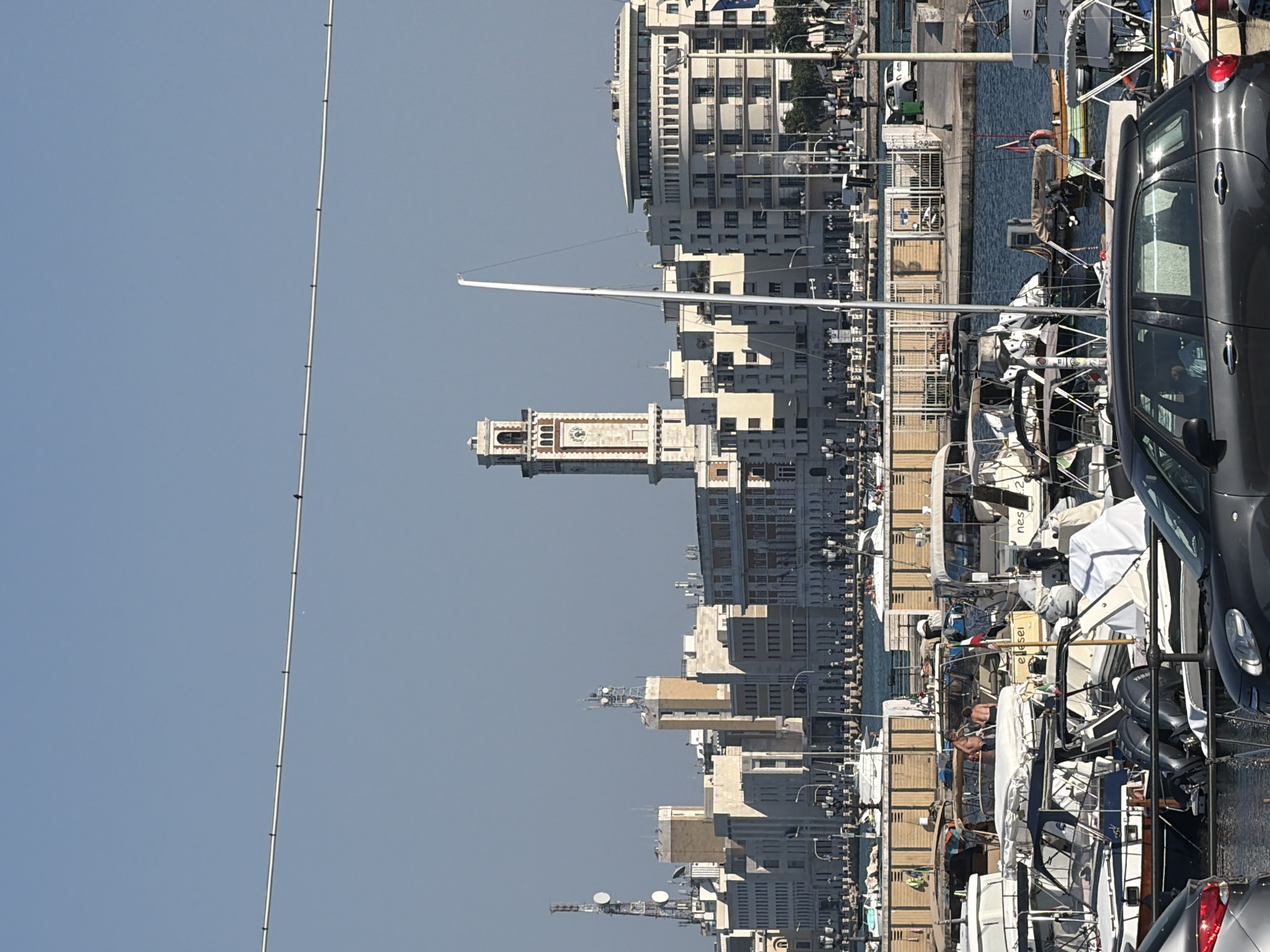 This is a daytime, eye-level, wide-angle photograph of the harbor in Bari, Italy, looking towards the city skyline. The foreground features a collection of docked boats of various sizes, some covered and some uncovered, with two figures in swimwear standing on the deck of one. A dark gray car is partially visible in the bottom left corner. The middle ground shows a waterfront promenade with lampposts and buildings, including what appears to be administrative or residential structures. Dominating the skyline in the middle distance is a prominent clock tower, characteristic of Italian city architecture. The background is a clear, light blue sky. The overall scene depicts a typical sunny day at a bustling Italian harbor.