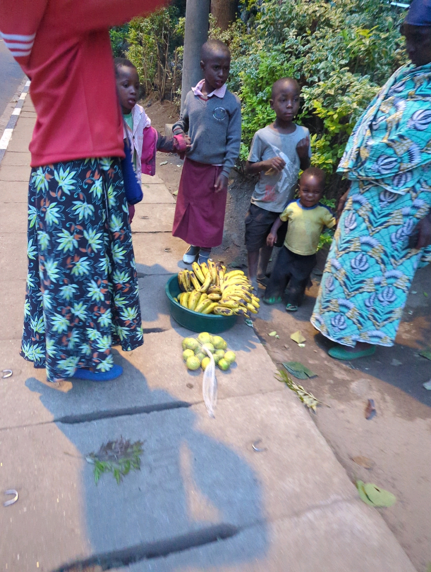 In Musanze District, Rwanda, a group of children and an adult are gathered on a sidewalk next to a road. The adult, in a brightly patterned blue and green dress, is standing to the right, partially obscuring a young child in a yellow shirt and black pants. To the left of the adult, a boy in a grey sweater and maroon skirt stands holding hands with a younger child wearing a pink and white striped shirt. Behind them, another boy in a grey t-shirt and dark shorts stands next to a large green bowl filled with bananas. A mesh bag of green citrus fruits sits in front of the bowl. The sidewalk is concrete, with visible cracks, and the background features dense green foliage and a utility pole. The scene appears to be outdoors, under natural lighting, with shadows cast across the ground.
