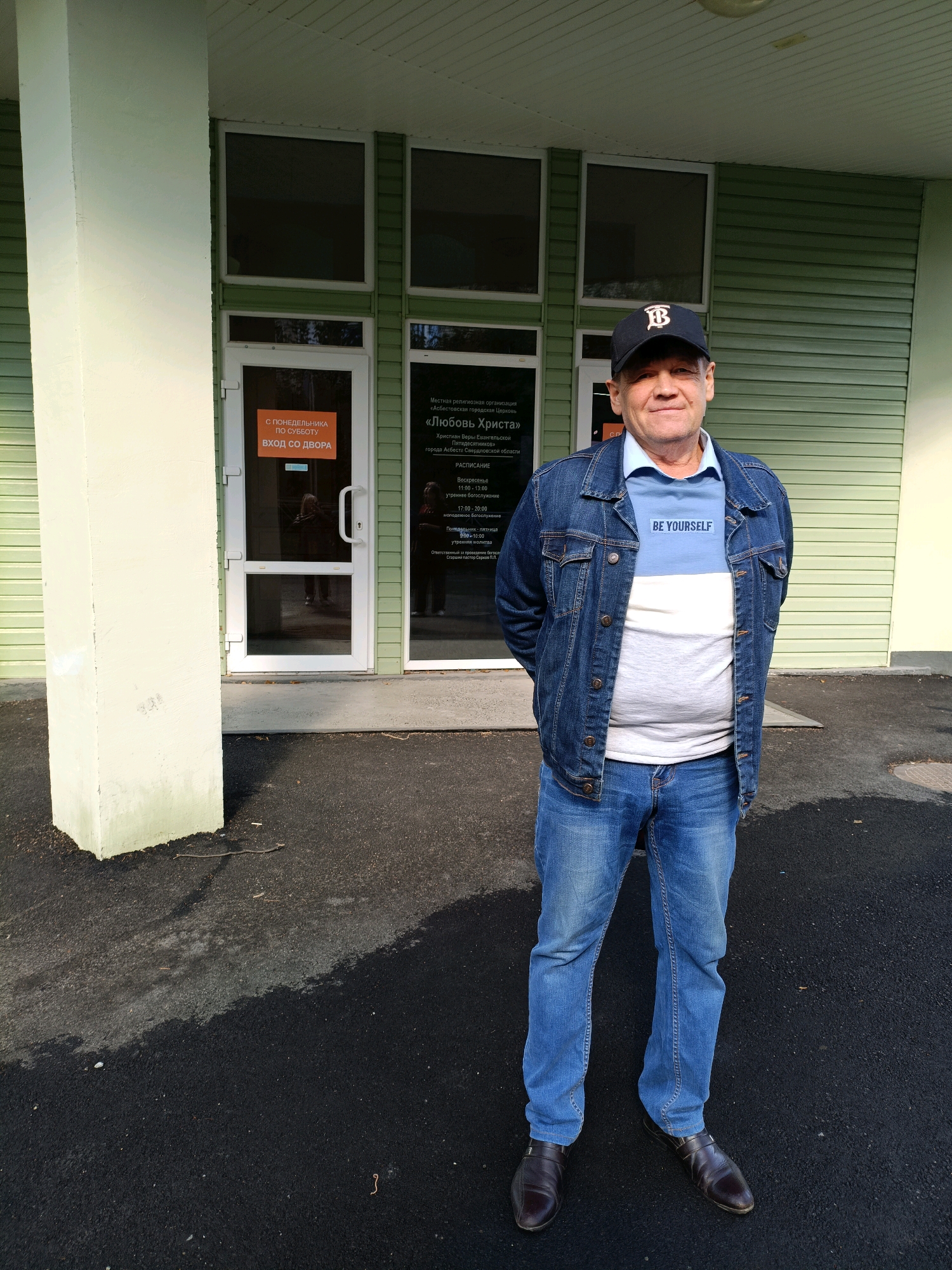 The image shows a middle-aged Caucasian man standing outside a building in Asbest, Russia. He is wearing a denim jacket, a light blue and white t-shirt with the text "BE YOURSELF" on it, blue jeans, and dark brown leather shoes. He has a black baseball cap with a white "B" logo on his head. The building appears to be a community center or a place of worship, with glass doors and windows. A sign on one of the doors reads "С ПОНЕДЕЛЬНИКА ПО СУББОТУ ВХОД СО ДВОРА," which translates to "FROM MONDAY TO SATURDAY, ENTRANCE FROM THE COURTYARD." Another sign on the adjacent door has text related to a religious organization. The ground in front of the building is paved with asphalt. The man is facing the camera with his hands behind his back, and he is smiling slightly. The lighting suggests it is daytime.