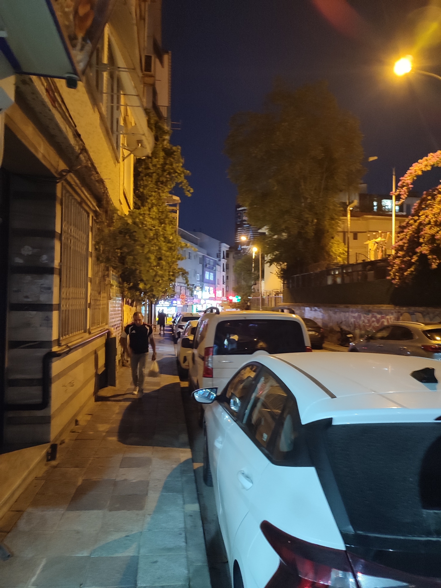 A man walks down a sidewalk in Kadıköy, Turkey, at night. He is wearing a dark shirt with a white stripe and light-colored pants. He carries a white plastic bag in his right hand. The sidewalk is made of gray and brown square tiles. To his left, there is a building with a storefront and barred windows. A tree with green leaves is growing next to the building. To his right, several cars are parked along the street. The streetlights cast a warm glow on the scene. The sky is dark blue, indicating it is nighttime. The scene appears to be a typical urban street with shops and parked vehicles.