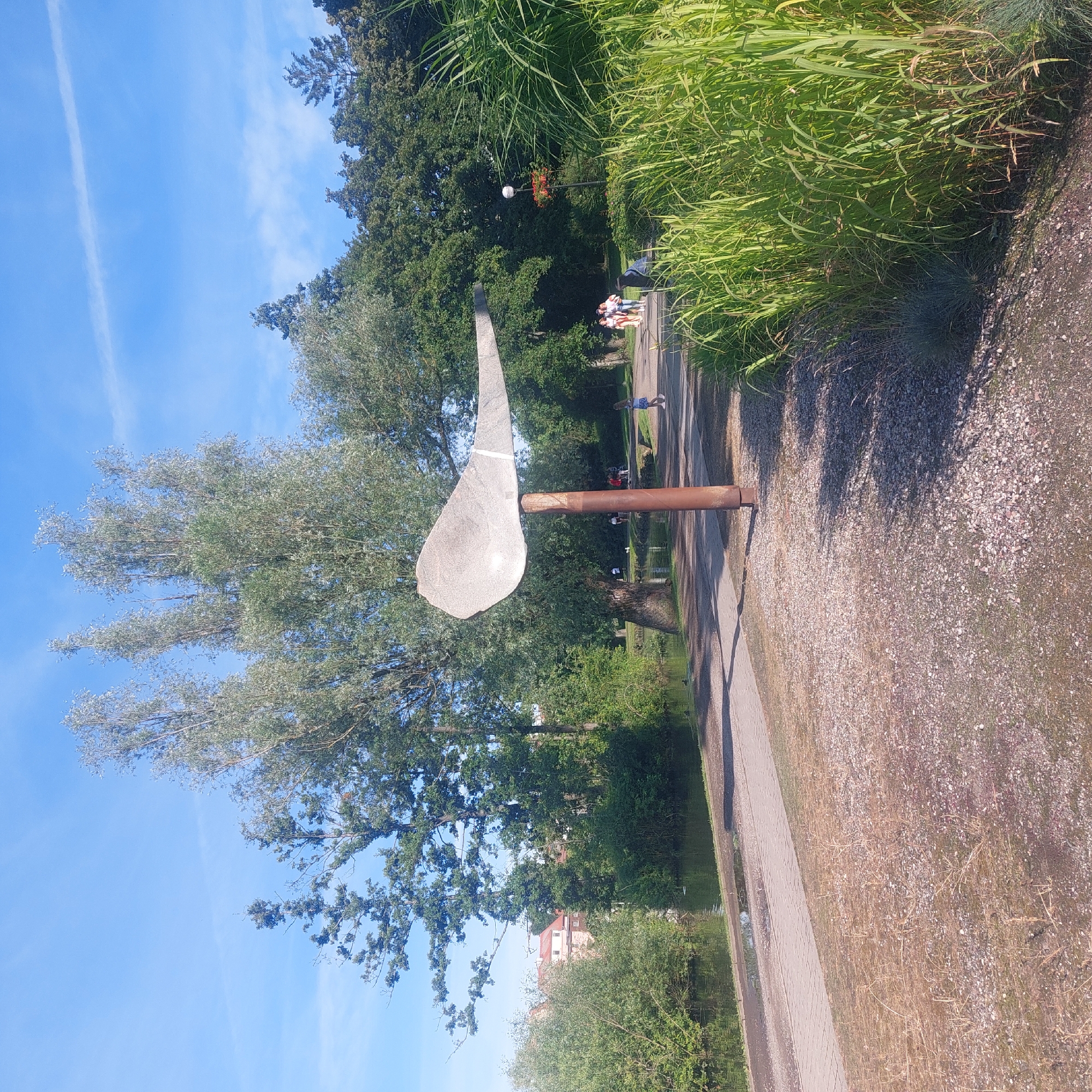The image depicts a sunny day in a park area in Mrągowo, Poland. In the foreground, a pathway made of stone slabs runs alongside a body of water on the left, bordered by green reeds on the right. A large, abstract sculpture resembling a bird or wing, made of light-colored material, stands on a rusty metal pole in the middle ground. Further back, a paved walking path curves through the park, with several people strolling along it. Trees and dense foliage form a backdrop to the scene, with a glimpse of buildings peeking through the greenery on the left. The sky is clear blue with a few wispy clouds.