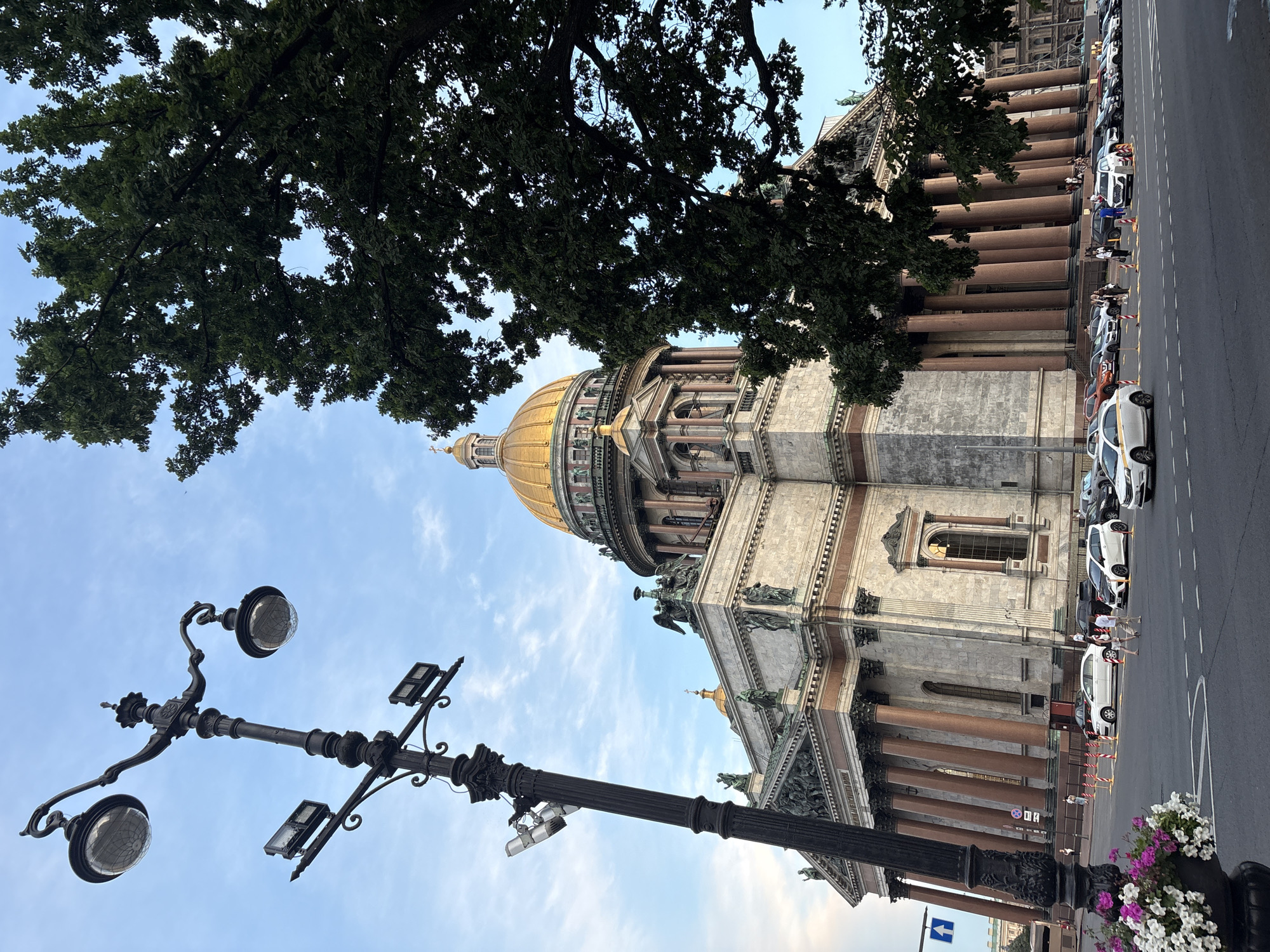 The image shows Saint Isaac's Cathedral in Saint Petersburg, Russia, viewed from a street level perspective. A large, ornate black lamppost is in the foreground on the left, leaning slightly. To the right and behind the lamppost, the majestic cathedral stands with its golden dome and classical architecture featuring numerous columns. A large tree with dense green foliage partially obscures the right side of the cathedral. Several cars are parked and driving on the street in front of the cathedral. A few pedestrians are visible crossing the street and walking near the building. The sky is partly cloudy, with a soft blue hue. In the lower left corner, a planter filled with white and pink flowers is partially visible. The scene appears to be captured during daylight hours, likely late afternoon or early evening, given the quality of light.