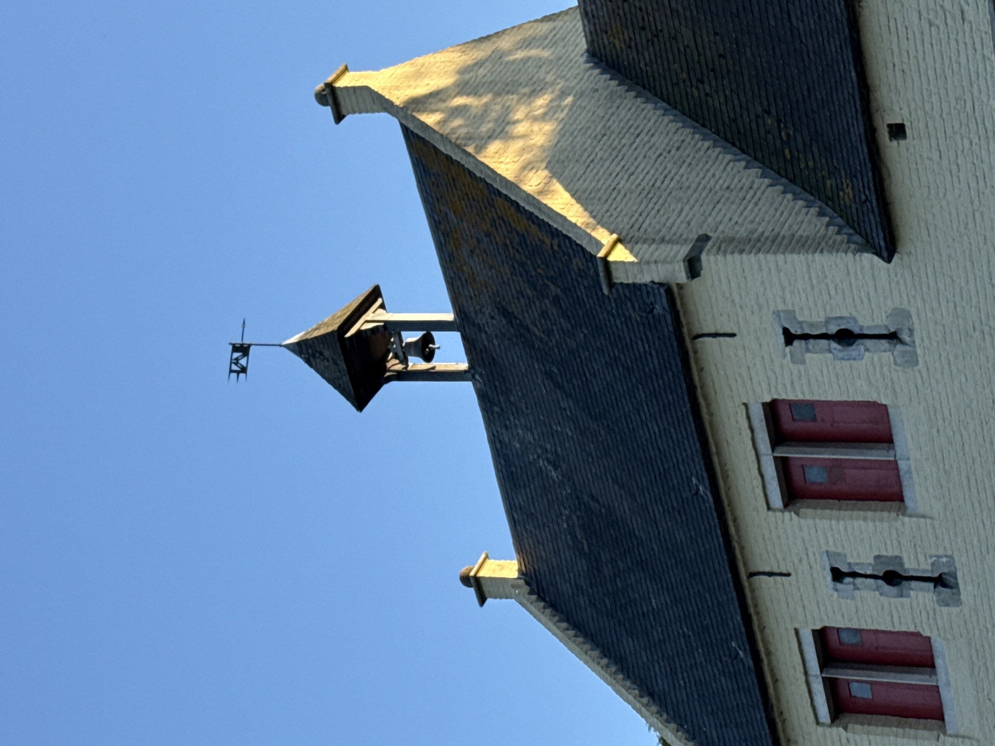 The image shows a low-angle view of a building's facade and roof against a clear blue sky. The building appears to be made of light-colored stone or stucco and features several windows with red shutters and decorative stone surrounds. The roof is covered in dark gray tiles and has a distinct architectural style with sharp angles and protruding decorative elements. At the apex of one roof section, there is a small bell tower with a weather vane. The sun is casting strong shadows on the roof, suggesting it is either morning or late afternoon. The scene is static, with no people or animals visible, and the overall impression is one of quiet architectural grandeur. The location context of Bruges, Belgium, suggests a historic European setting, possibly a church, guild hall, or other public building.