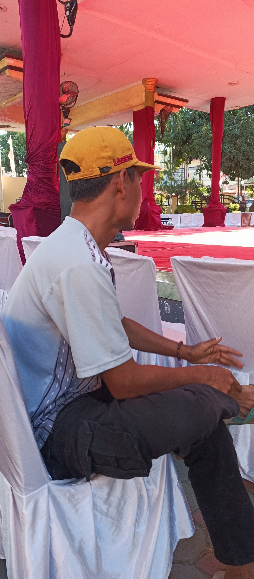 A man wearing a yellow cap with the word "LOGISTIC" on it is sitting on a white-covered chair, facing to the right. He is wearing a light-colored t-shirt with a gray and white patterned design on the sleeves and side. His pants are dark gray and appear to be cargo-style. His right hand is raised, palm open, in a gesturing motion. His left arm rests on his lap, and his legs are crossed. Behind him, there is a red carpeted stage area with red draped fabric and golden pillars. Trees and buildings are visible in the background. The scene appears to be outdoors, possibly at an event or gathering in Arjasa, Indonesia.