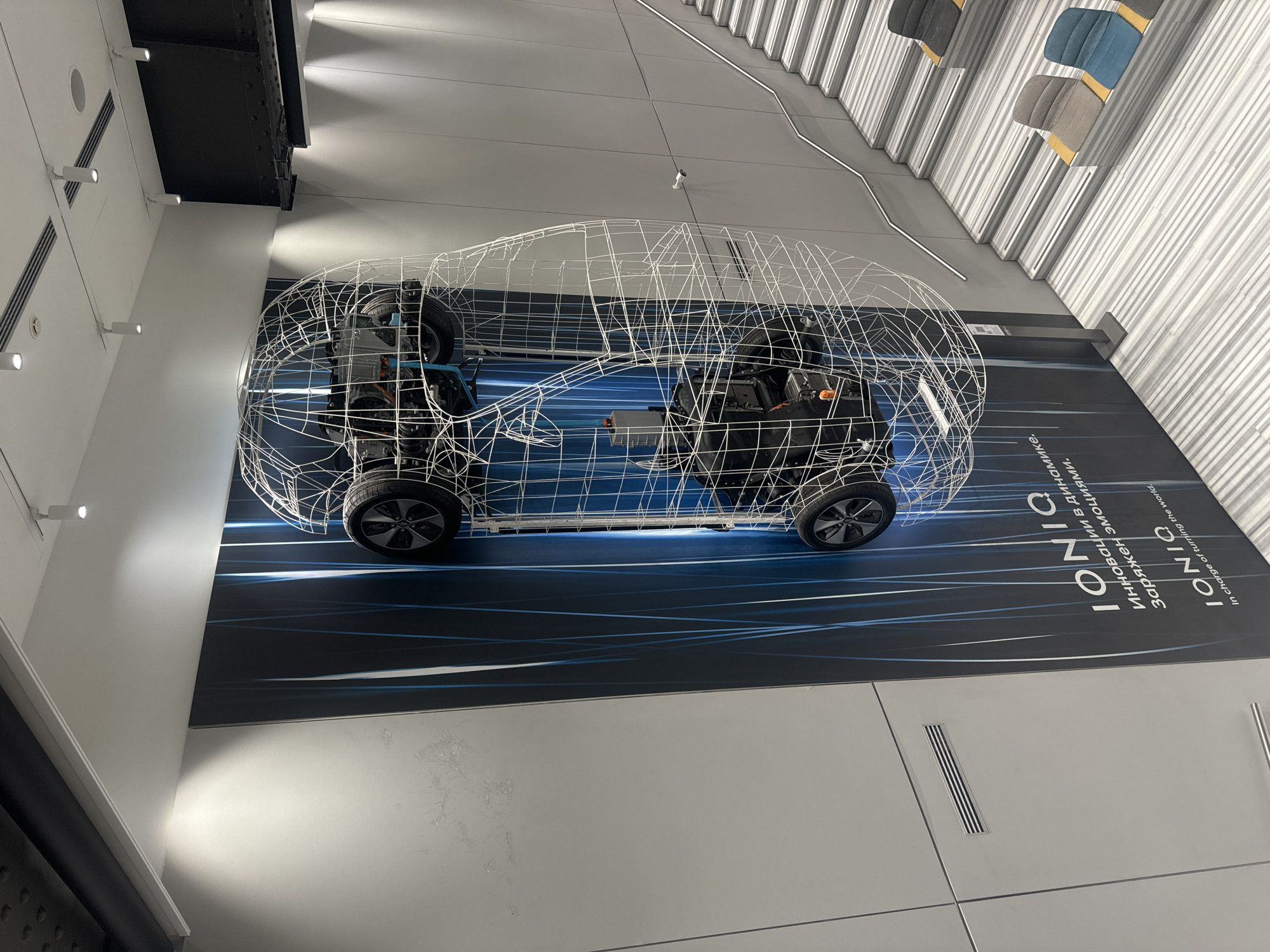 The image captures a modern indoor space, possibly a showroom or exhibition hall, showcasing a skeletal wireframe model of a car, specifically the Hyundai IONIQ 5. The model sits on a display panel featuring branding and informational text, including the car's name and tagline in both English and Russian. The overall aesthetic is sleek and contemporary, with the architecture featuring clean lines and a minimalist color palette of whites, grays, and blacks. The lighting is bright and evenly distributed, accentuating the car model and the surrounding environment. Based on the writing, and the overall modern styling, it is plausible that this location may be in Moscow, Russia.
