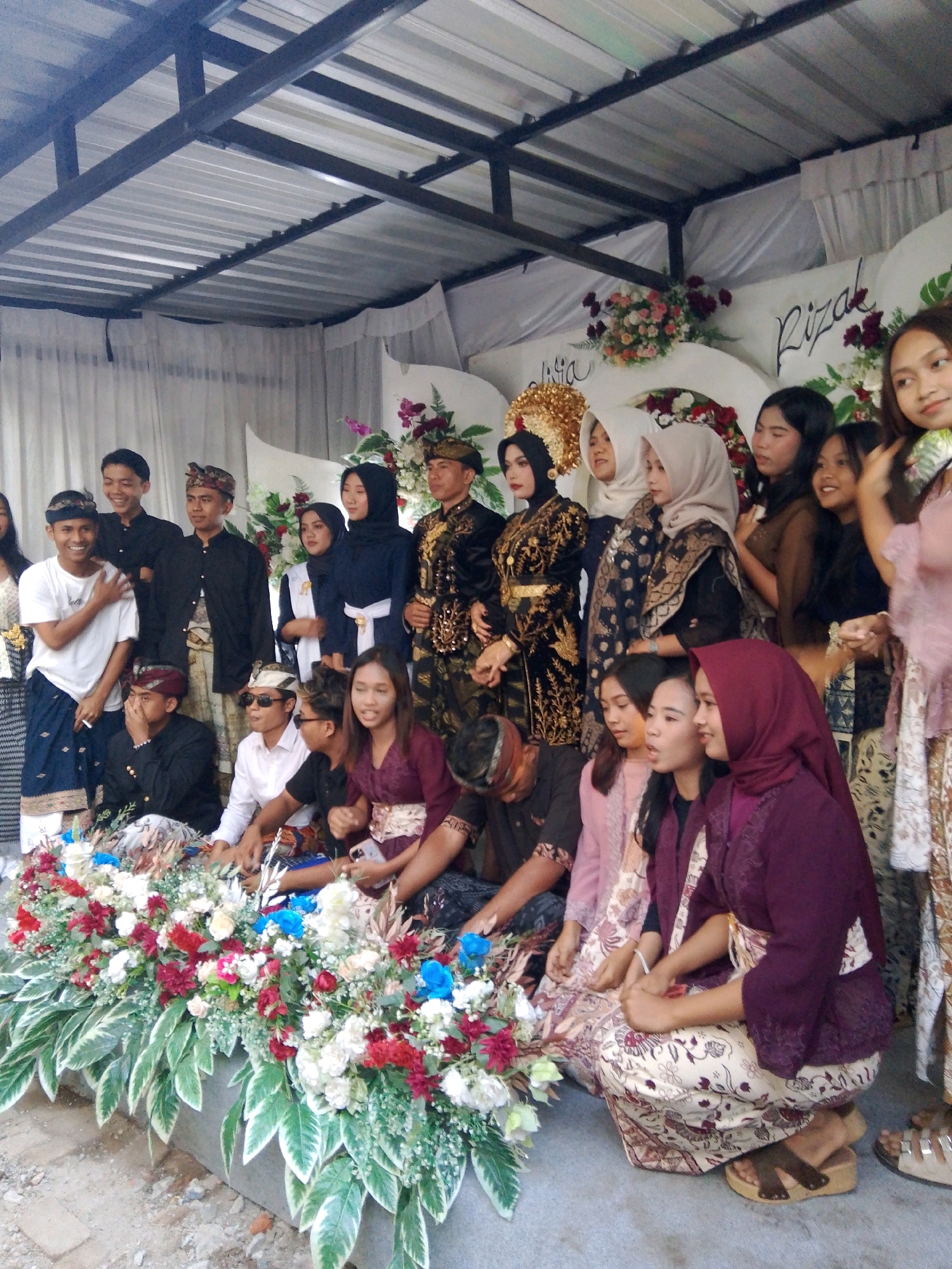 The image shows a group of people, likely attending a ceremony or celebration, gathered in what appears to be an outdoor or semi-outdoor setting with a corrugated metal roof overhead. The individuals are dressed in traditional attire, including batik patterns and headscarves. Several people are wearing formal clothing, while others are in more casual traditional wear.
In the foreground, there is a large floral arrangement with a variety of flowers in red, white, and blue, along with green foliage. Some people are seated on the ground near the floral arrangement.
In the background, there is a white backdrop with some decorative elements and a handwritten name that appears to read "Rizal." The overall atmosphere suggests a cultural event. The image is taken from a slightly elevated perspective, looking down at the group. The lighting is natural, suggesting it was taken during the daytime. The ground appears to be concrete or packed dirt. There are no identifiable landmarks of Kuripan Utara, Indonesia, in the image.