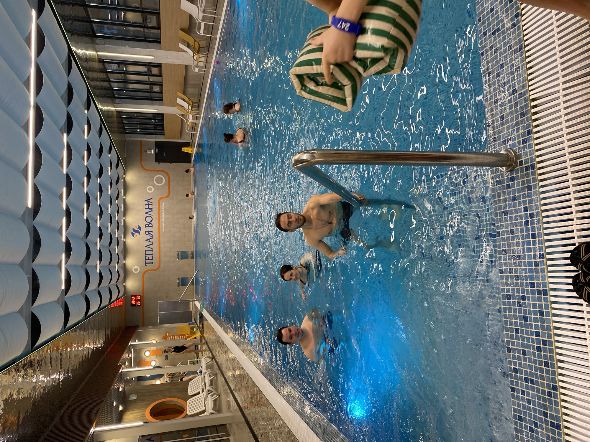 The image shows an indoor swimming pool in Votkinsk, Russia. The pool is filled with blue water and several people are swimming and standing in it. A man is holding onto a stainless steel pool ladder. Another man is smiling at the camera. A person holds a green and white striped bag. The pool's edge is lined with blue and white mosaic tiles. The building features a wavy pattern on the ceiling, and windows are visible along the wall opposite. A sign on the wall reads "Теплая Волна" in Cyrillic. The time is displayed on a digital clock as 19:42.