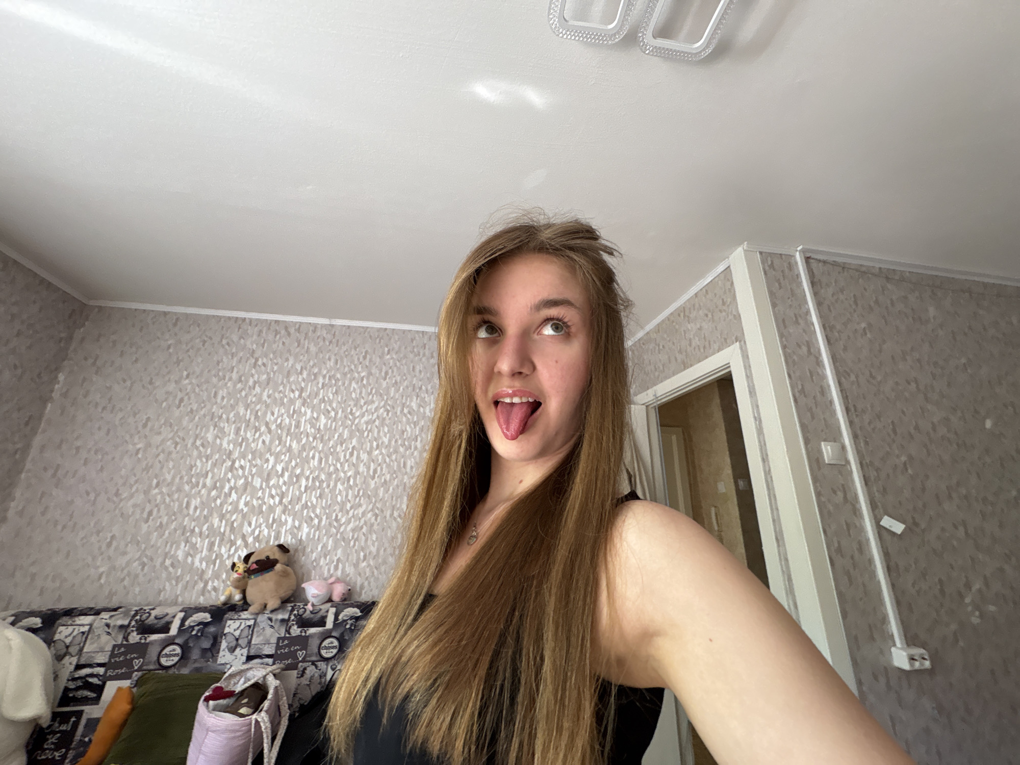 A young woman is holding the camera up high and looking upward with her tongue out. She has long, straight, light brown hair and is wearing a black top. The room appears to be inside an apartment. There is a couch with patterned upholstery in the background, with several stuffed animals on it. The walls have textured wallpaper. A doorway is visible in the background, as well as a light fixture on the ceiling. Based on the details of the interior and location metadata, the image was likely taken inside an apartment in the city of Perm, Russia.