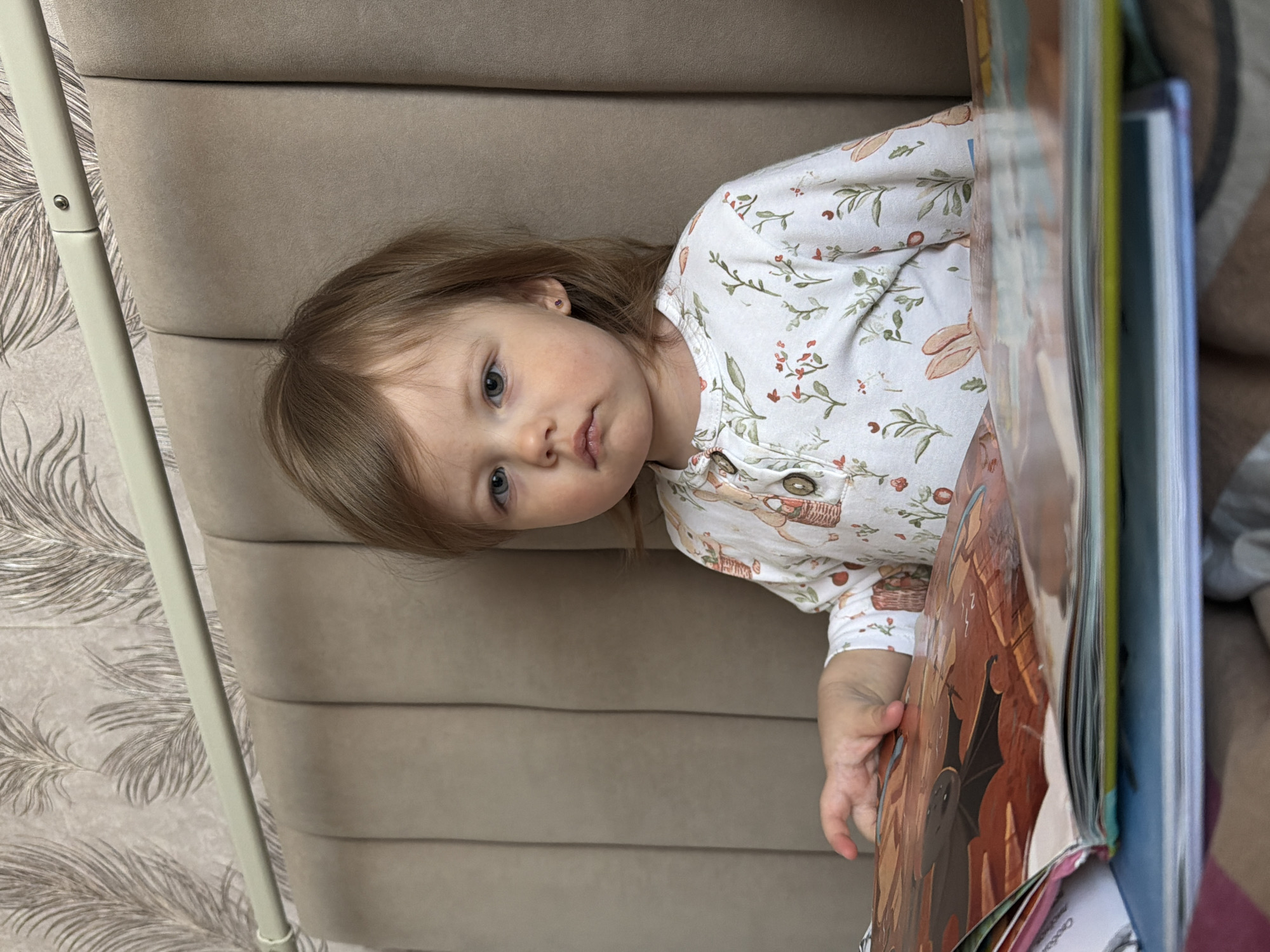 The image shows a young child, likely a toddler, lying on their side against a cushioned headboard or wall. The child is wearing a light-colored, long-sleeved garment patterned with floral and basket motifs. The child is partially obscured by a stack of books, one of which is open, revealing an image of what appears to be autumn leaves and a bat.
It's impossible to verify the location of Reshetnikovo, Russia, or any other geographic location without additional information.