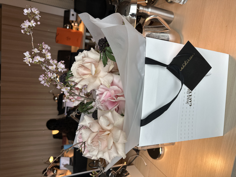 A bouquet of light pink and cream roses, delicate pink cherry blossoms on branches, and dark spiky foliage, wrapped in white tissue paper, rests inside a white paper gift bag with black fabric handles. The bag is branded "jean paul GAULTIER" and has a black rectangular tag displaying "grandiflora" in cursive script attached to one handle. This arrangement is centered on a light wooden desk within a modern office environment in London, United Kingdom, indicated by the "grandiflora" tag, which is a known London-based florist. In the blurred background, two individuals are seated at separate workstations, each equipped with a computer monitor and a silver desk lamp emitting warm light. An orange gift bag is visible on a distant desk, and a reflective silver pitcher or shaker set is partially visible to the immediate right of the floral arrangement. The far background features light-toned vertical wall panels or blinds.