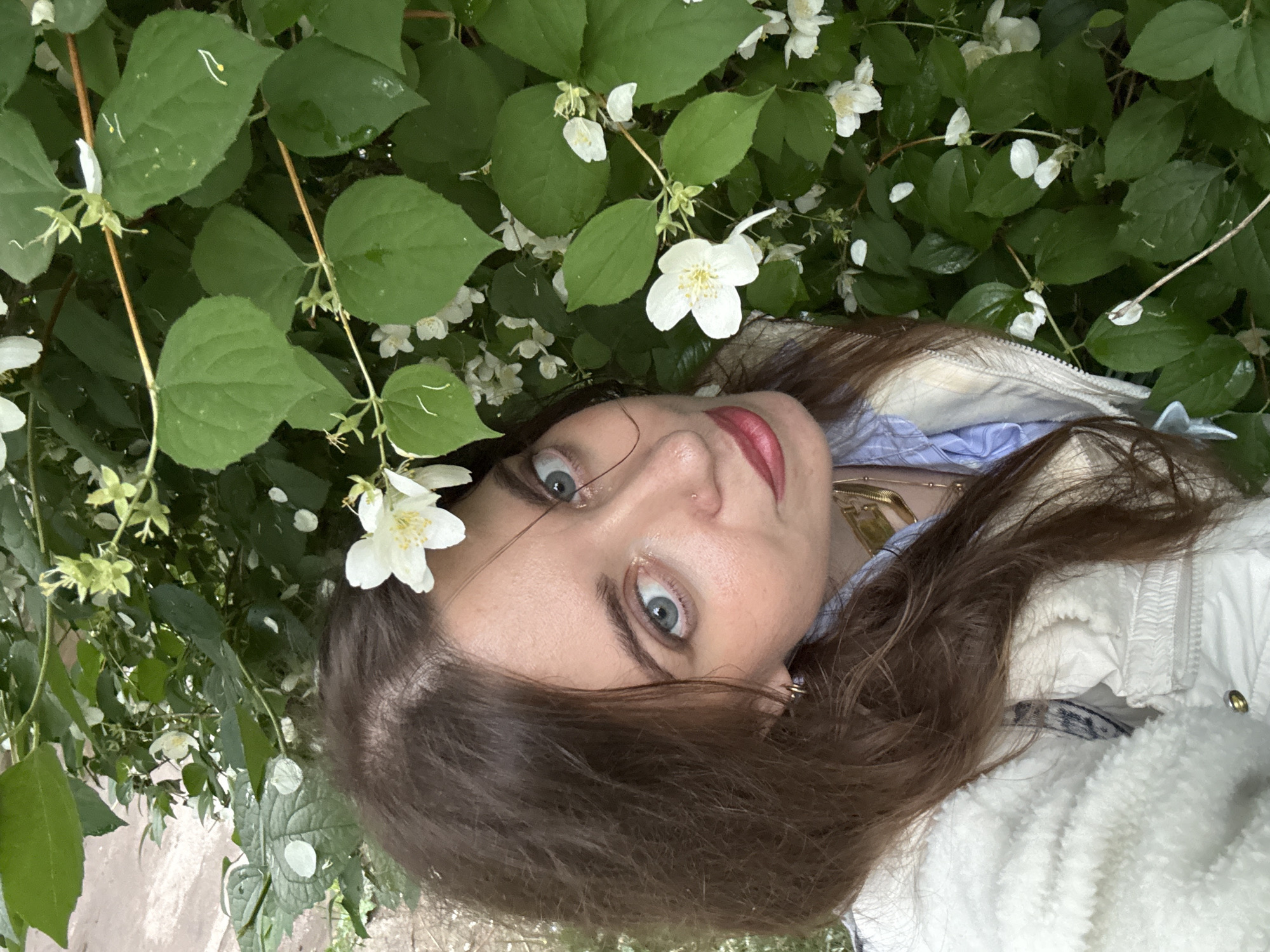 A close-up, overhead shot features a young woman lying amongst lush green foliage and white flowers. Her eyes, wide and blue with flecks of green, are directed towards the camera, framed by long brown hair. Her lips are painted a vibrant red, and a small nose ring is visible. She wears a light-colored jacket with a fluffy white collar over a blue shirt and a delicate gold necklace. One white flower rests near her left eye, and other blossoms are scattered throughout the leaves. The scene appears to be outdoors, possibly in a garden, with natural lighting suggesting daytime. The overall mood is serene and slightly whimsical, with the woman seemingly relaxed and engaged with the natural surroundings. There is no visible text in the image.