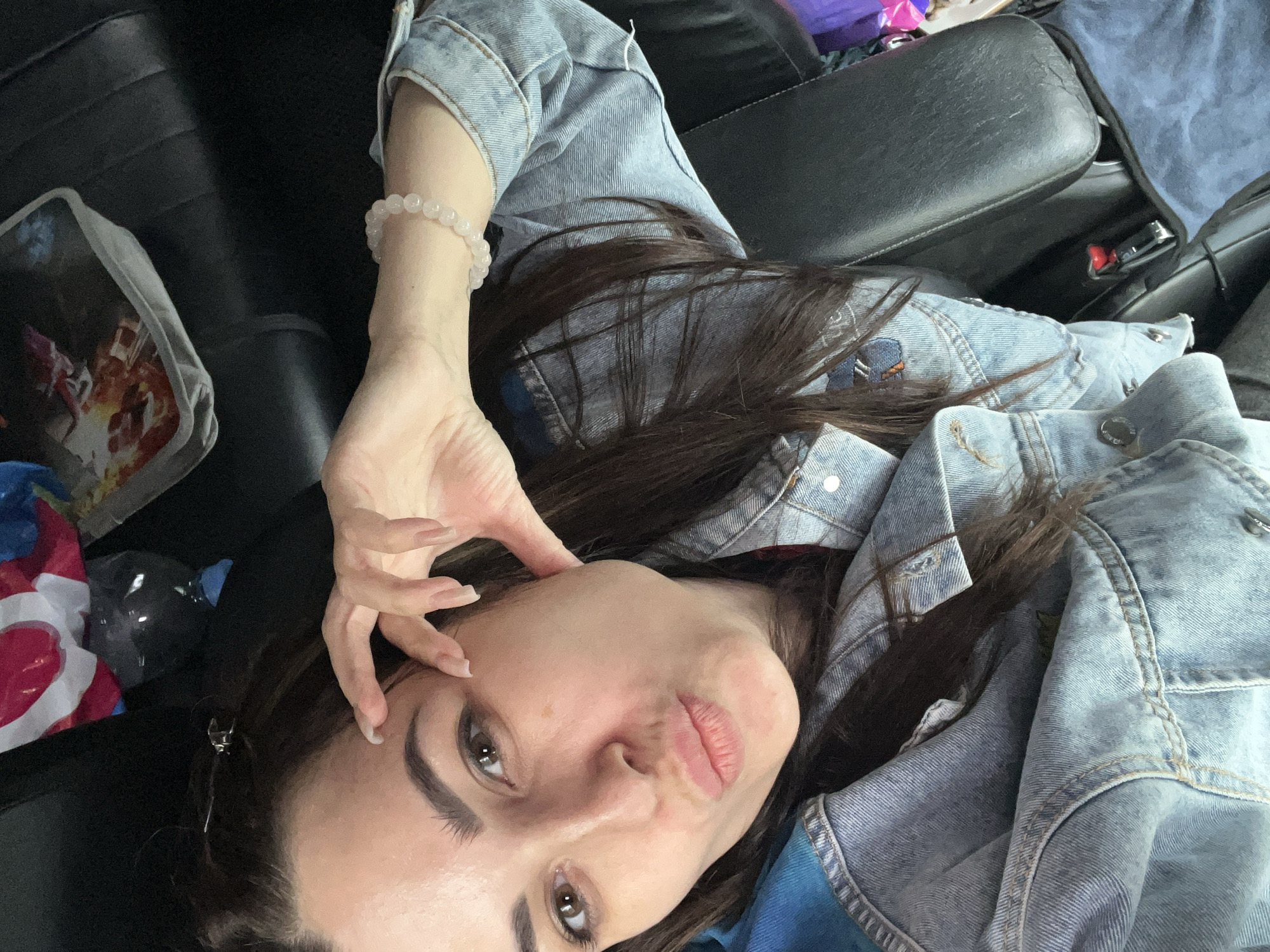 A woman is lying down. She is wearing a light blue denim jacket and has a bead bracelet on her wrist. Her dark hair is spread around her. She is looking at the camera with a slightly puckered expression. In the background, there is a black leather surface, possibly a car seat. There is a light-colored bag to the left of the woman. The location is Unknown, Unknown.