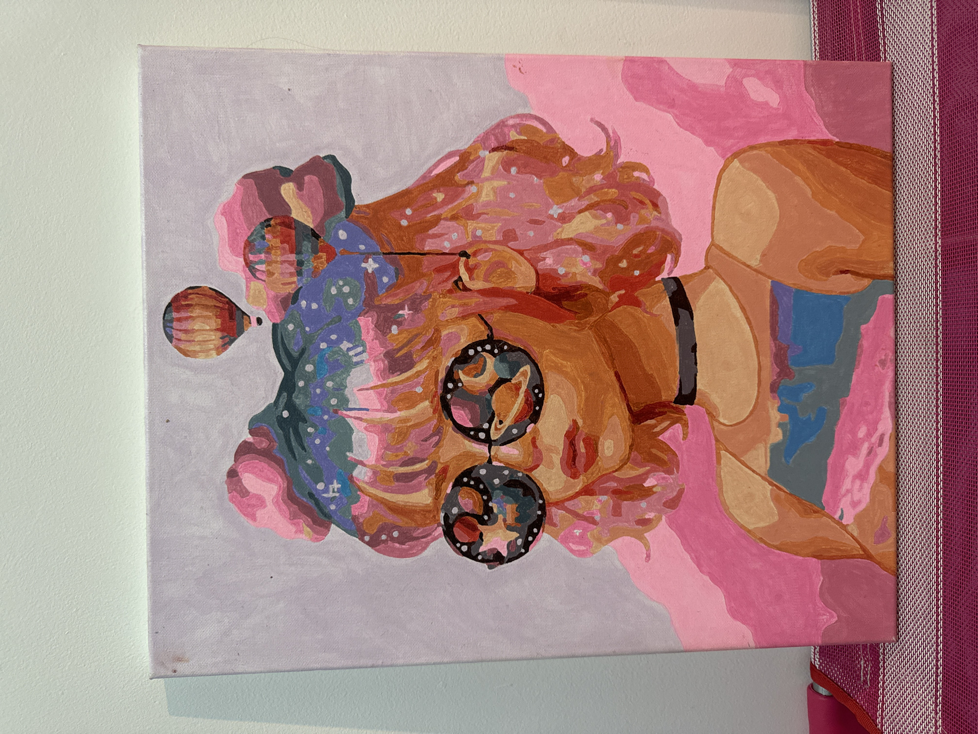 A vibrant, stylized painting depicts a person, likely a woman, from the chest up in profile, facing right. The subject has voluminous, curly hair rendered in orange, peach, pink, and yellow tones, dotted with white and light-colored specks. Large, circular earrings, intricately detailed with celestial bodies like planets and stars, dangle from their ear. A dark, choker-style necklace adorns the neck. A prominent, patterned hot air balloon shape appears near the back of the head, possibly an elaborate headdress or an abstract element. The skin is portrayed with flat planes of orange and peach.
The painting is a portrait, so no specific activity or event is depicted.
The art style is graphic and contemporary, utilizing distinct block-color areas with minimal blending. The color palette is warm and rich, featuring prominent pinks, oranges, and purples, contrasted with cooler blues and greys. The background is abstract, with a soft lavender/grey section on the left and fluid shapes in various shades of pink on the right. The artwork is executed on a canvas.
No visible text is present in the painting. While provided in the context of Mũi Né, Vietnam, the artwork itself does not contain explicit geographical cues for this location.