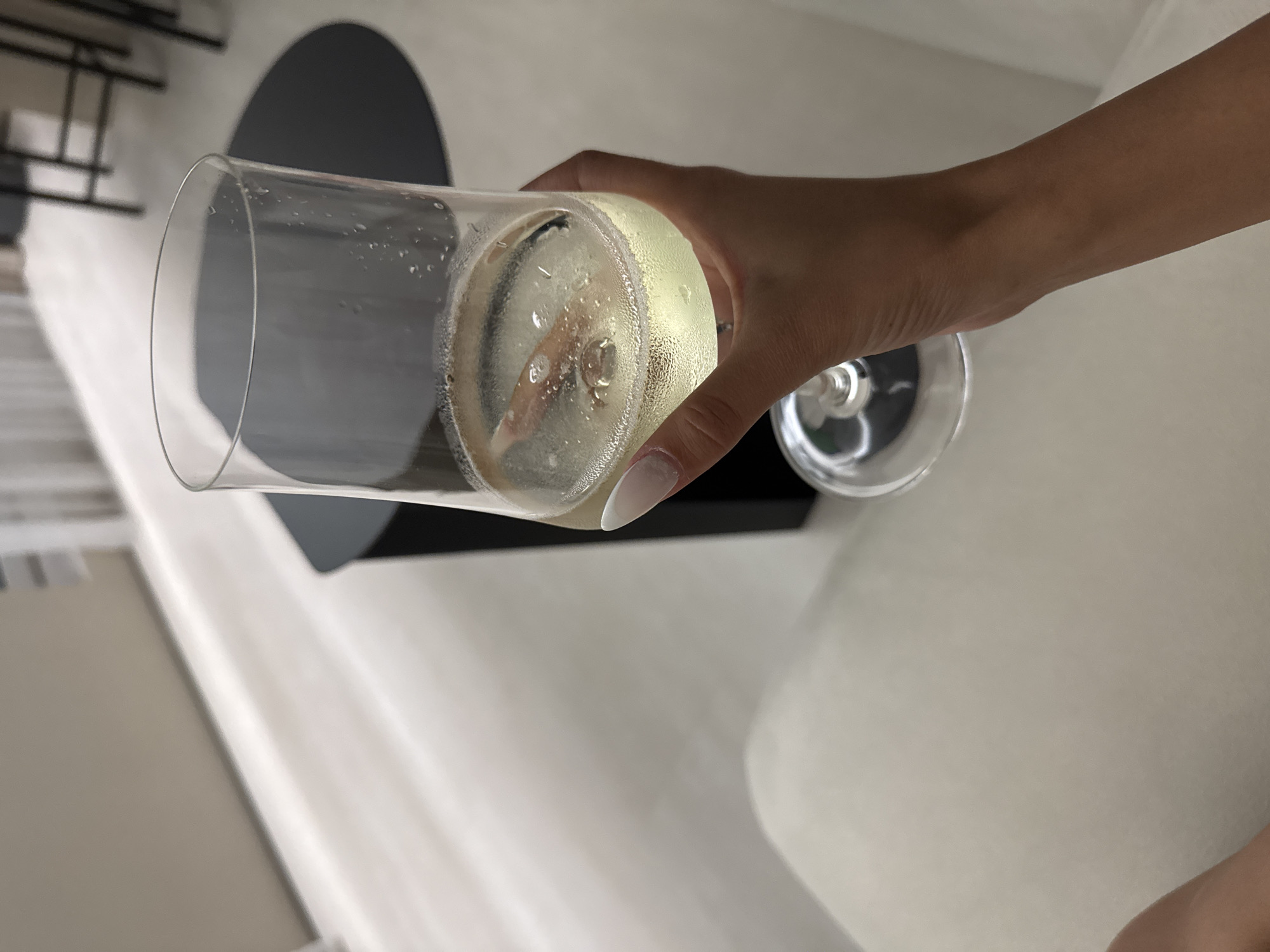 A person's hand with long, manicured nails holds a tall, clear glass filled with a light-colored, effervescent beverage. The glass has condensation on its exterior and contains what appears to be a cinnamon stick submerged in the liquid. The background is softly lit and appears to be an indoor setting with a light-colored floor and furniture. There are dark, cylindrical objects visible in the background, possibly stools or table legs. The scene does not provide any specific visual cues that would place it in the city of Izhevsk, Russia.