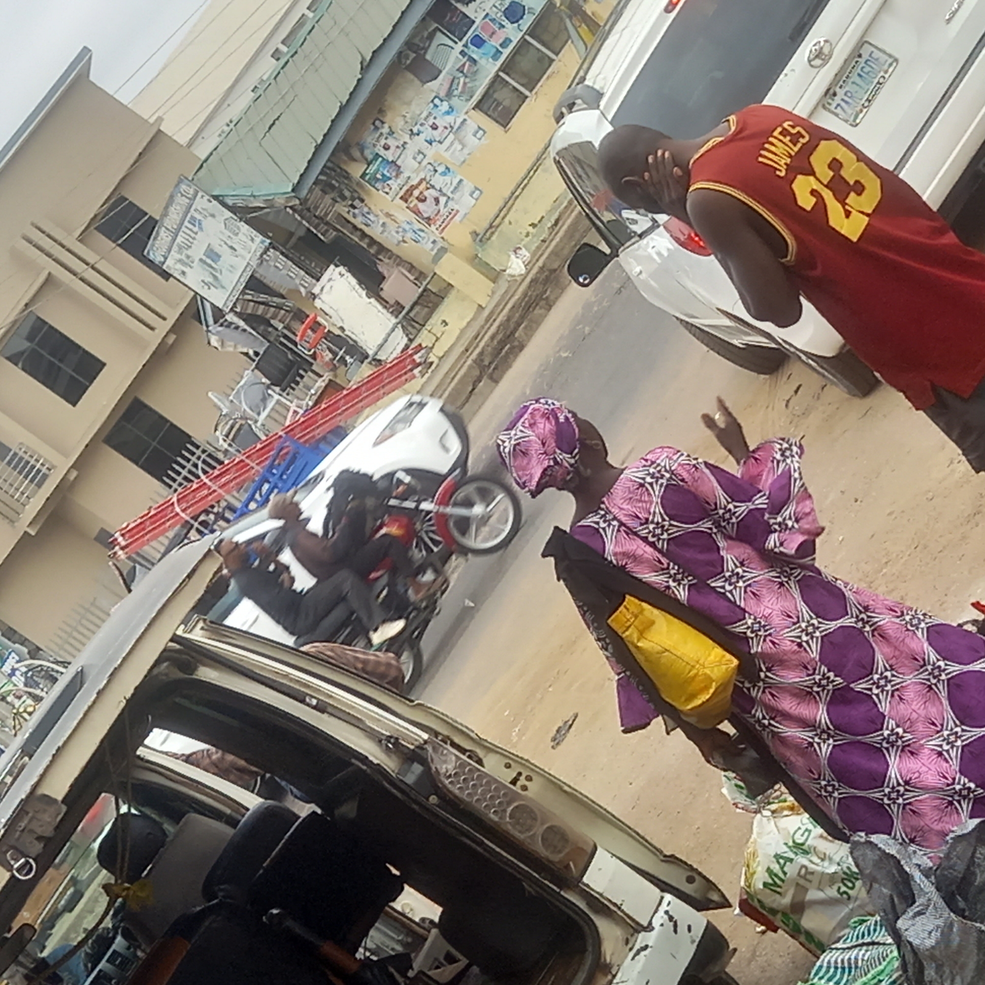 The image shows a street scene in what appears to be an urban area of Dadi Riba, Nigeria. An accident has occurred involving a car, a motorcycle, and possibly another vehicle. The car is tilted and has its interior exposed. The motorcycle lies on the ground with a rider on top of the car. A red ladder is visible, possibly cargo from one of the vehicles. Two people are standing near the scene: one wearing a purple patterned garment and carrying a yellow bag, and another wearing a red basketball jersey with the name "JAMES" and the number "23" on the back. The person in the jersey has their hands on their head. Another white car is visible on the street with a license plate starting with "ZAR-14". The street has some garbage bags next to the purple dressed person. There is a building with faded posters and a shop sign in the background.