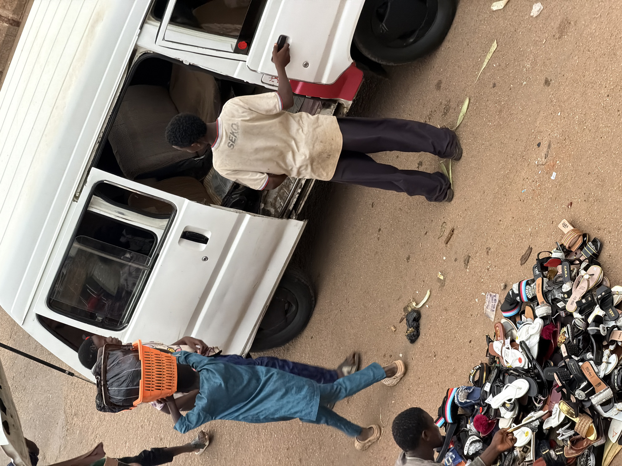 In Bukuru, Nigeria, a street scene unfolds with a white minibus, its doors open. A young boy, dressed in a blue tunic and pants, carries a black bag in an orange basket on his head, walking towards the minibus. Another boy, in a beige t-shirt with "SEKO" printed on it and dark pants, stands near the open side door of the minibus, his hand on the door frame. A pile of assorted footwear, including sandals and flip-flops, is visible on the dusty ground near the foreground, with a person's hands interacting with the pile. The environment suggests a bustling roadside market or transport hub.