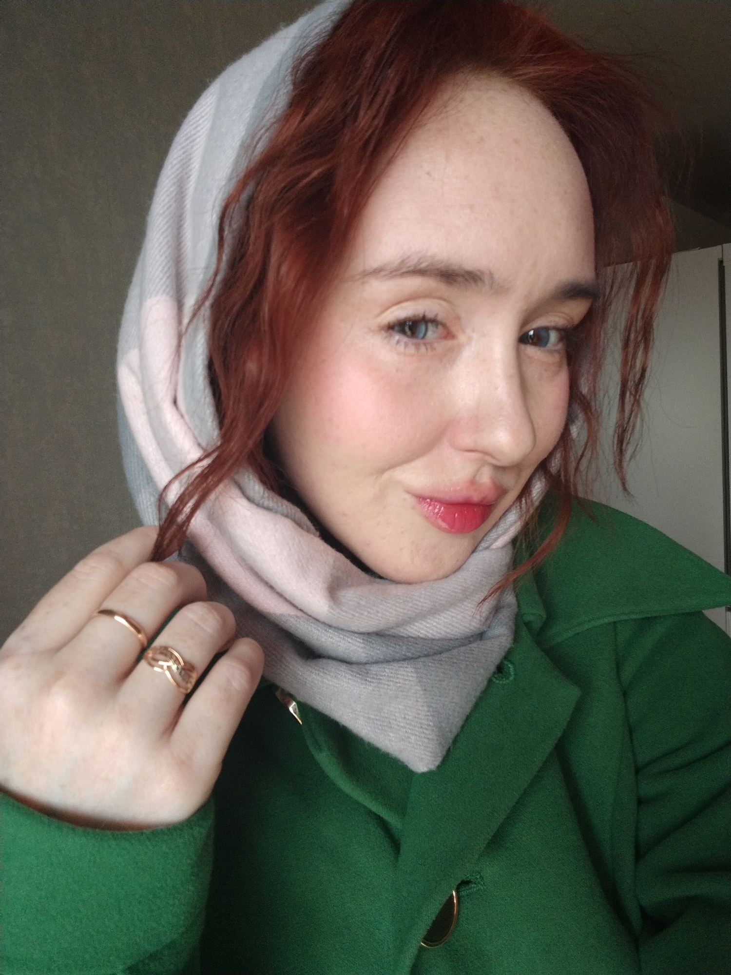 A woman with red hair and blue eyes is posing indoors. She is wearing a green coat and a light grey and pink scarf that covers her head. Two gold rings are visible on her left hand. The background is neutral, with a wall and a door partially visible. There is no clear indication of Izhevsk, Russia.