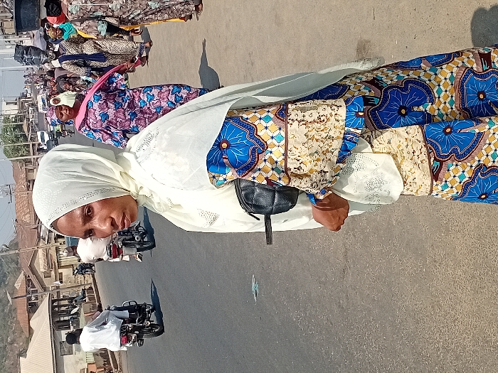 A woman wearing a white hijab and colorful dress stands on a street. Other women in similarly vibrant attire are nearby. Motorbikes and buildings line the background. The scene suggests a bustling outdoor environment, likely in a sunny location.