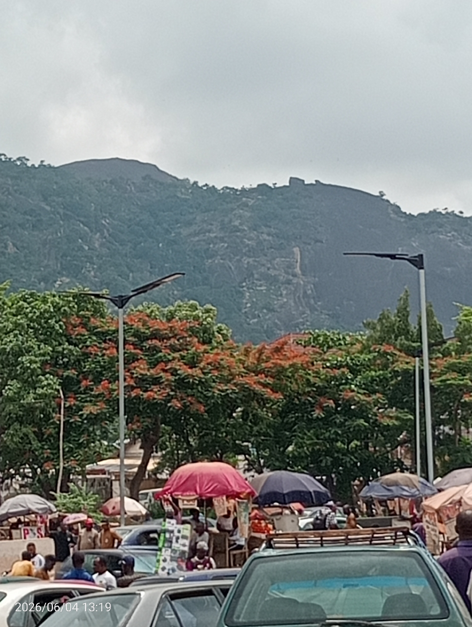 The image depicts a bustling street and market scene in Madalla, Nigeria, set against a backdrop of verdant mountains under an overcast sky.
In the foreground and mid-ground, numerous people are gathered amidst cars, suggesting a lively market or roadside commerce. Several cars are visible, including a silver sedan, a green vehicle with a roof rack, and others partially obscured. Market stalls, characterized by a variety of colorful umbrellas in shades of red, blue, brown, and white, are spread across the area, indicating places where vendors sell goods. Individuals are seen standing, walking, and interacting, with some appearing to engage in transactions.
Prominent in the mid-ground are several large trees, some of which feature striking clusters of orange-red blossoms, likely Flame Trees (Delonix regia), contrasting with the dominant green foliage. Tall streetlights with distinctive, flat, wide luminaires (possibly solar-powered) stand tall, extending above the tree line.
The background is dominated by large, rolling mountains or hills, heavily covered in green vegetation, particularly noticeable in the upper half of the frame. The sky is uniformly grey and cloudy, indicating overcast weather conditions.
A digital timestamp "2026/06/04 13:19" is visible in the lower-left corner, dating the image to early afternoon. A partial white sign displaying "P S" can be seen near one of the market stalls. The overall scene conveys a sense of vibrant community activity integrated with the natural landscape.