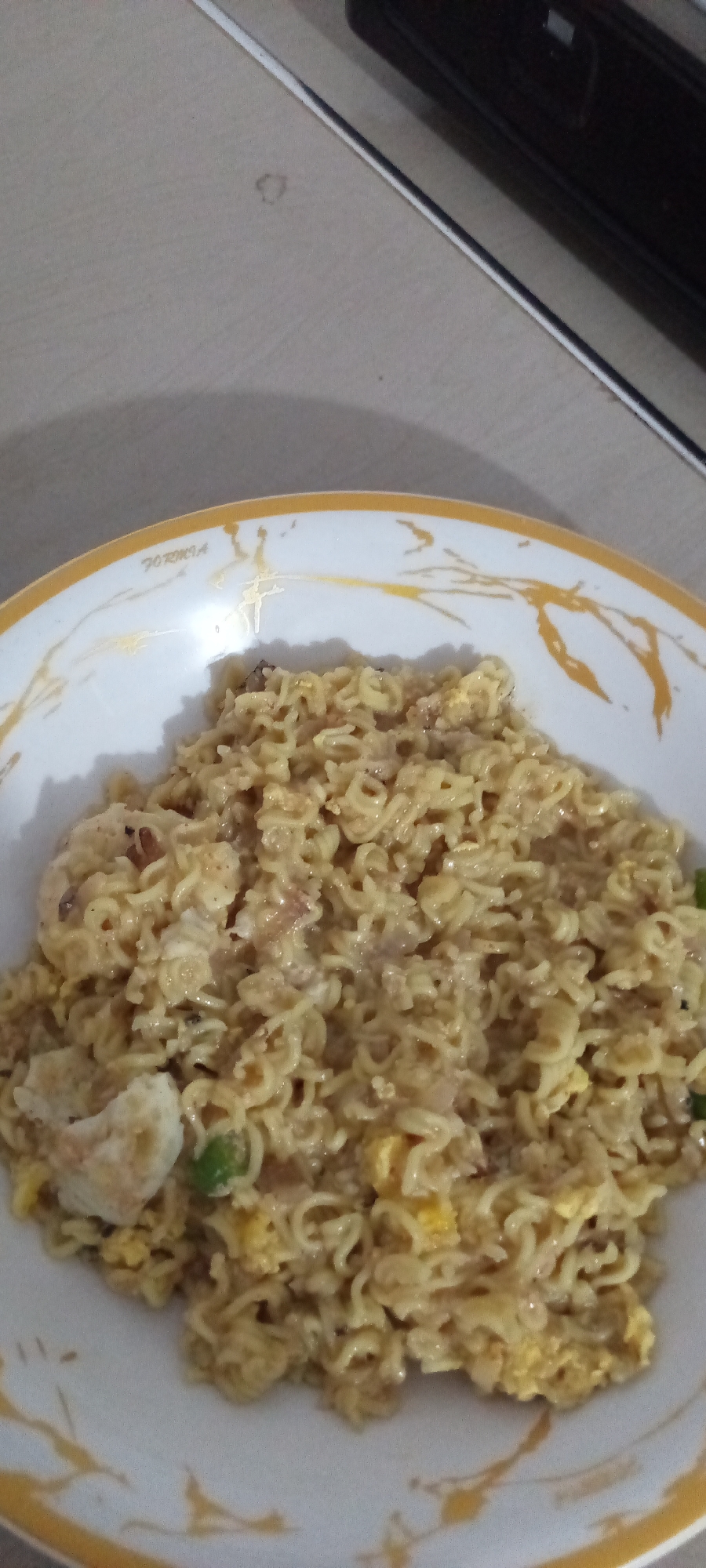 A close-up overhead shot shows a bowl of instant noodles mixed with scrambled eggs, onions, and possibly some chili pepper, on a tiled countertop. The bowl is white with a gold rim and a decorative gold pattern. The noodles are a light yellow-brown color and appear to be slightly wet. The eggs are visible throughout the noodles, appearing as yellow and white pieces. A few green specks, likely chili, are also scattered throughout the dish. The background consists of a neutral-colored tiled surface and the edge of a black appliance, possibly a microwave or oven. The lighting is soft and appears to be natural, casting some shadows. The scene is set in Lhokseumawe, Indonesia.