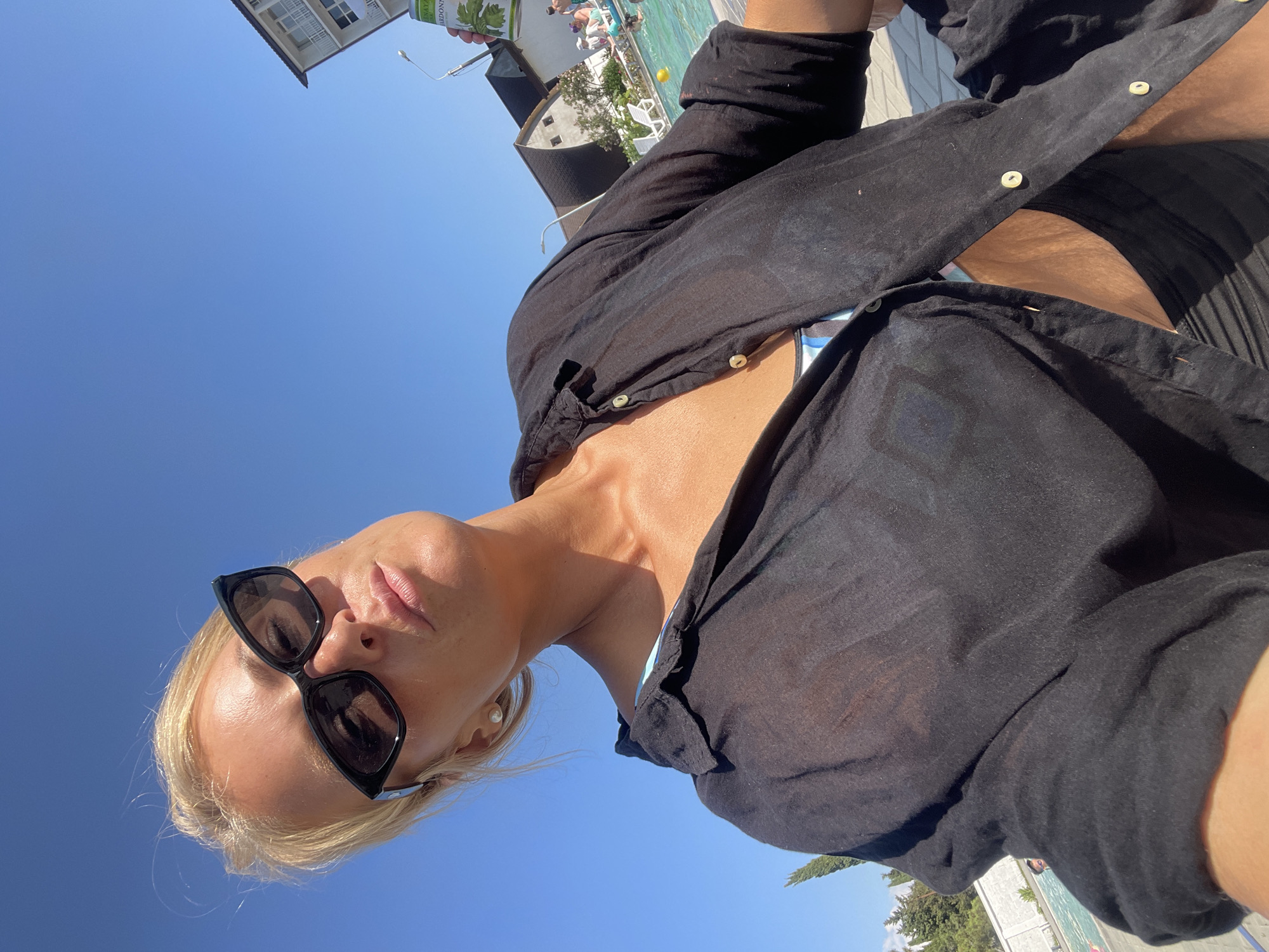 A woman with blonde hair and sunglasses takes a selfie outdoors, leaning back and looking at the camera. She is wearing a black open shirt over a bikini and black shorts. Behind her, a swimming pool with lounge chairs and people can be seen, along with some buildings and trees. The sky is clear and blue. The scene appears to be a resort or hotel in Giachrypsh, Abkhazia.