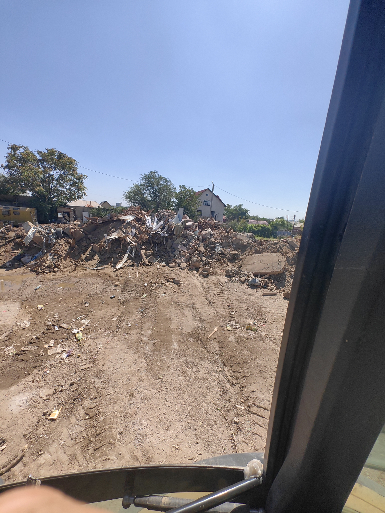 The image depicts a demolition site in Shymkent, Kazakhstan, characterized by a large pile of rubble, including concrete, metal, and other building materials. In the background, a multi-story building with a white facade and dark windows stands amidst trees. The ground is dirt with tire tracks, suggesting recent activity by heavy machinery. Scattered debris, such as plastic bottles and wrappers, is visible on the ground. The sky is clear and blue. The perspective suggests the photo was taken from within a vehicle, likely an excavator or similar heavy equipment, with a portion of the vehicle's interior visible in the foreground.