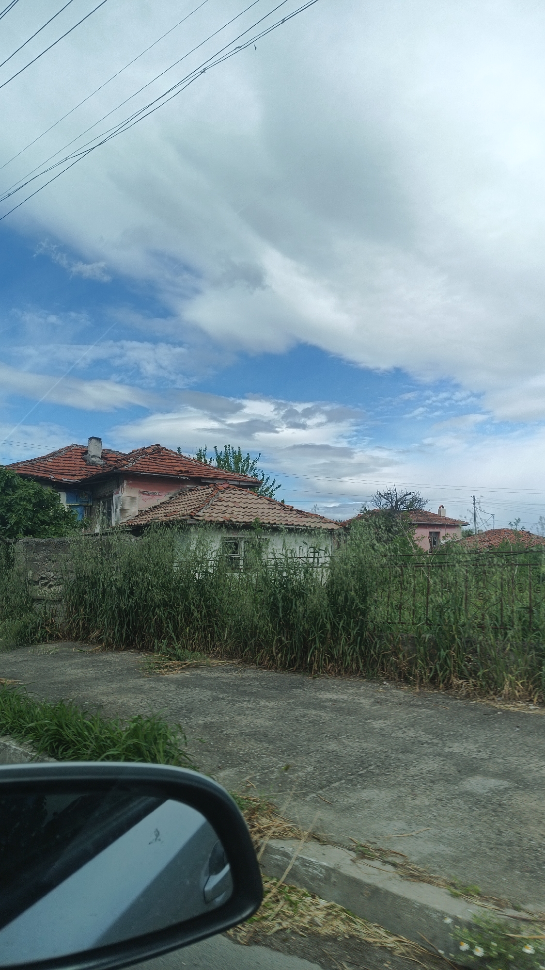 The image shows a view from a car in Milevo, Bulgaria. The car's side mirror is visible in the lower left corner. The road is concrete and there is an overgrown verge at the roadside. Beyond the verge are several old houses with red tile roofs, partially obscured by overgrown vegetation. The houses appear to be in disrepair. The sky is cloudy with patches of blue. Overhead power lines are visible against the sky.