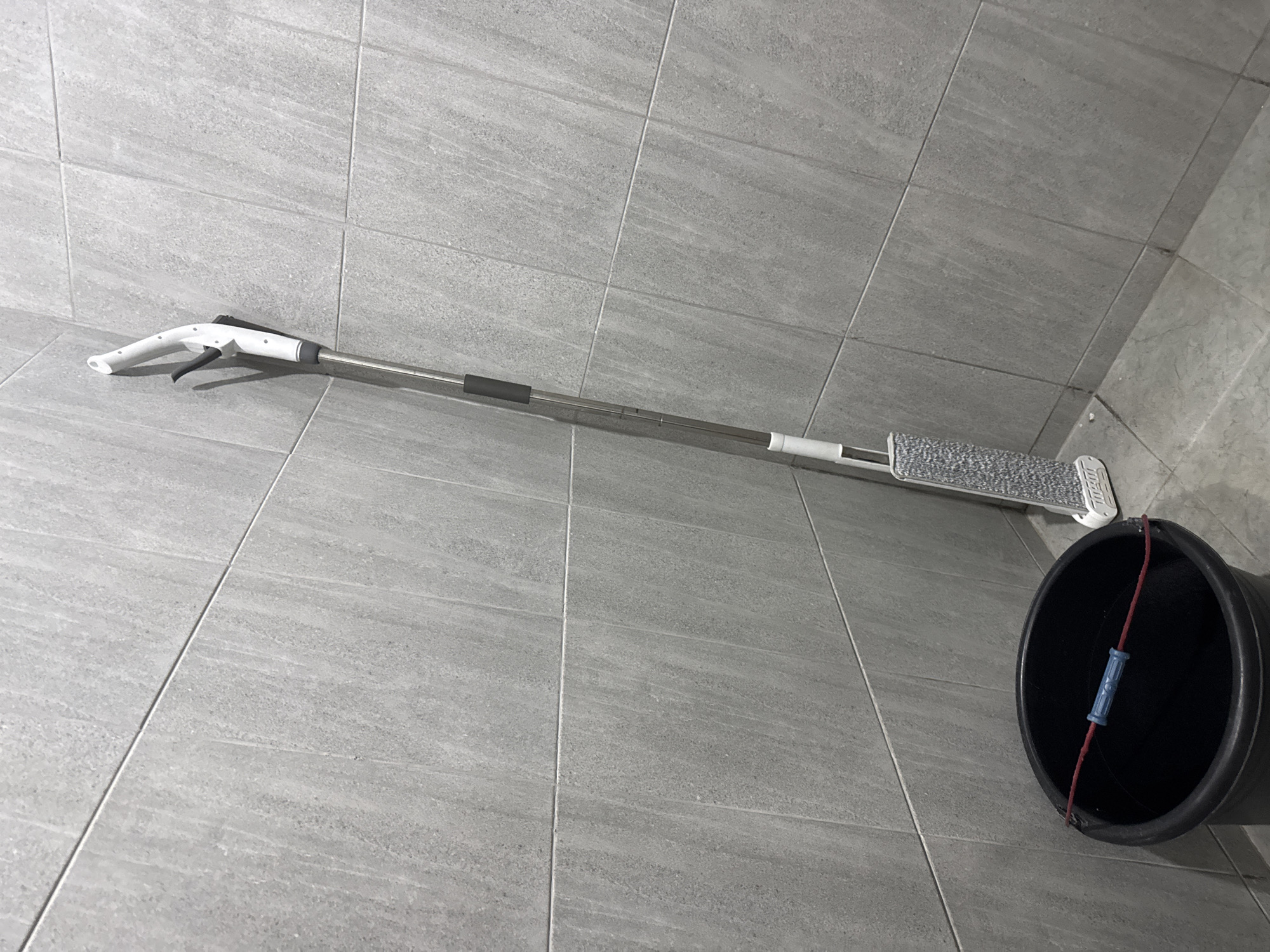 In a corner of a room with grey tiled walls and floor, a black bucket with a red rope and blue handle is placed on the floor. Next to the bucket, a flat mop with a grey microfiber pad stands upright. The mop has a silver handle with a black grip and a white dispenser at the top. The scene appears to be in a bathroom or utility area. There are no people present. The location is not specified in the image, and therefore cannot be determined to be in Sukawati, Indonesia.