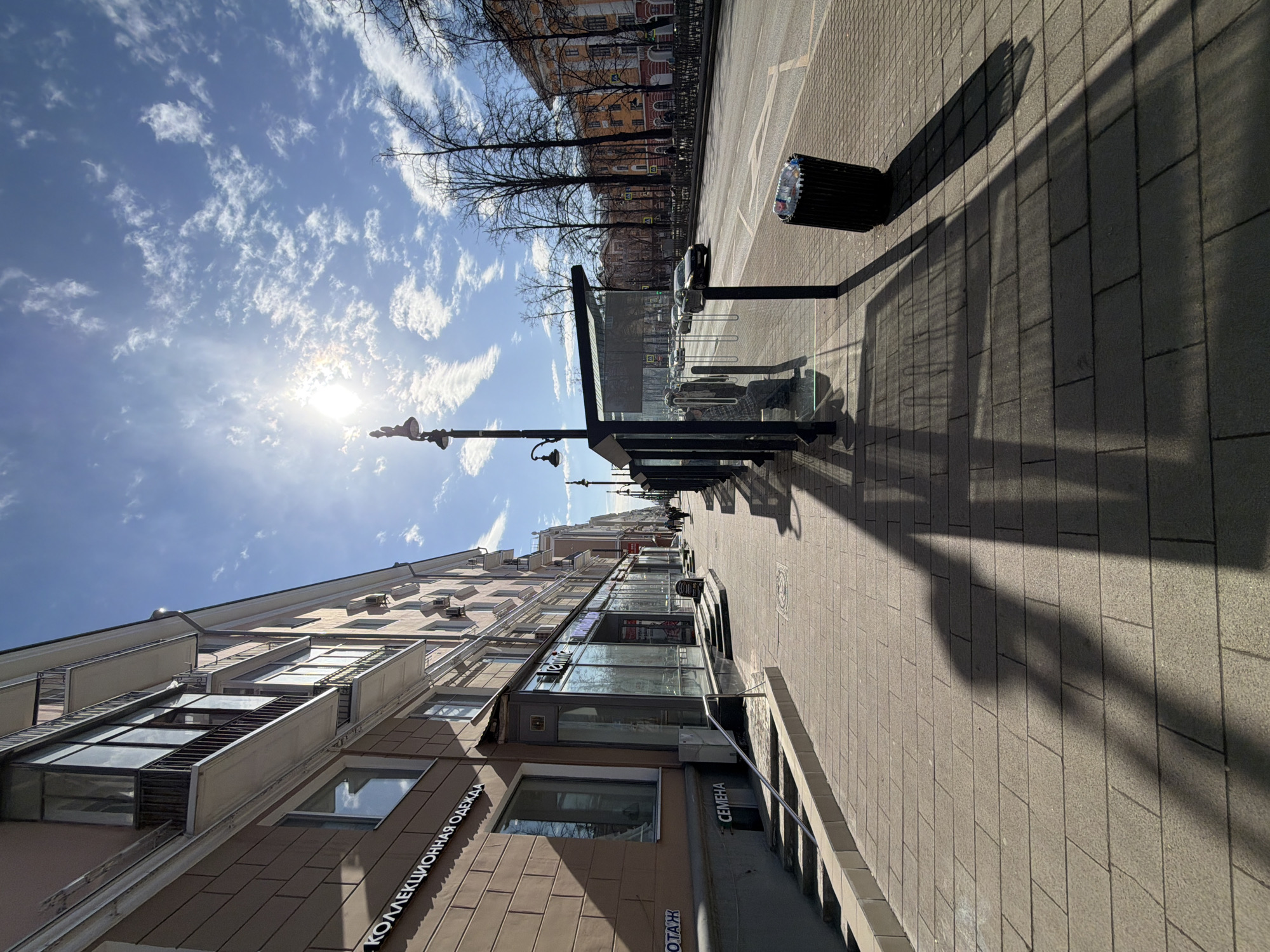 This image depicts a sunlit urban street scene on Komsomolsky Prospekt, specifically near building number 49, in Perm, Russia. The perspective is from a wide, light-colored paved sidewalk, looking eastward down the street.
To the immediate left, a multi-story building with a light brown-pink facade features large ground-level storefront windows and upper-floor balconies. Russian text "КОЛЛЕКЦИОННАЯ ОДЕЖДА" (Collection Clothing) and "СЕМЕНА" (Seeds) are visible on its exterior. Further down this sidewalk, several indistinct figures are observed walking.
In the mid-ground on the right side of the sidewalk, a modern bus stop shelter with a dark frame and glass panels is present. One person is seated within the shelter, partially obscured by shadows, appearing to wear a plaid garment. Another individual stands near the shelter. A black cylindrical trash receptacle, containing a clear plastic bottle and other refuse, is positioned next to the bus stop. Multiple ornate dark streetlights line the sidewalk.
Long, sharp shadows from the streetlights, the bus stop, and other unseen elements stretch across the light-colored paving blocks, indicating a low sun angle from the right (south).
Beyond the sidewalk, a multi-lane asphalt road with painted markings, including a dedicated bus lane, accommodates several vehicles, primarily cars, moving in both directions. A dark-colored sedan is visible in the middle distance.
Across the road, another multi-story building with a warm yellow-orange facade is visible, fronted by a row of tall, bare deciduous trees. Distant figures can be seen walking on the far sidewalk.
The sky is bright blue with scattered white cumulus clouds. The sun, a prominent bright disc causing lens flare, is positioned in the upper-left quadrant of the frame. The overall impression is a clear, cool, and sunny day, likely in late autumn, winter, or early spring given the bare trees.
