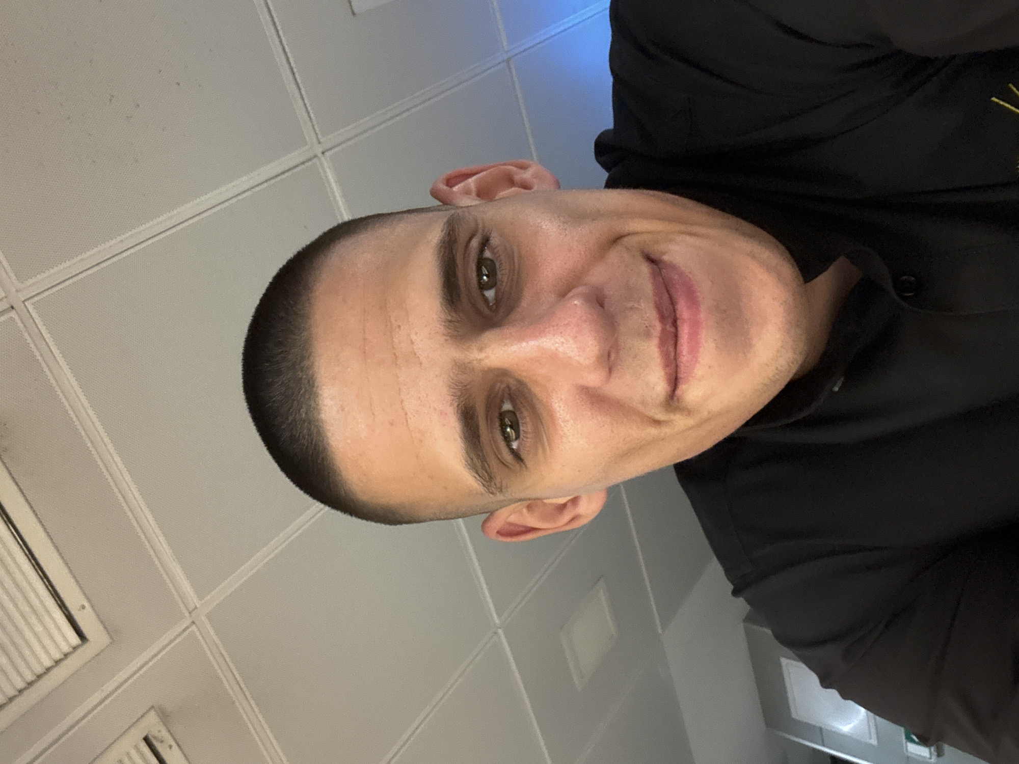 A close-up, selfie-style shot shows a man with a buzz cut and light brown eyes. He is looking directly at the camera with a slight smile. He is wearing a black collared shirt. The background appears to be an office or indoor space with a tiled ceiling. There is a blue light source visible in the upper right corner. The image was taken in Sofia, Bulgaria.
