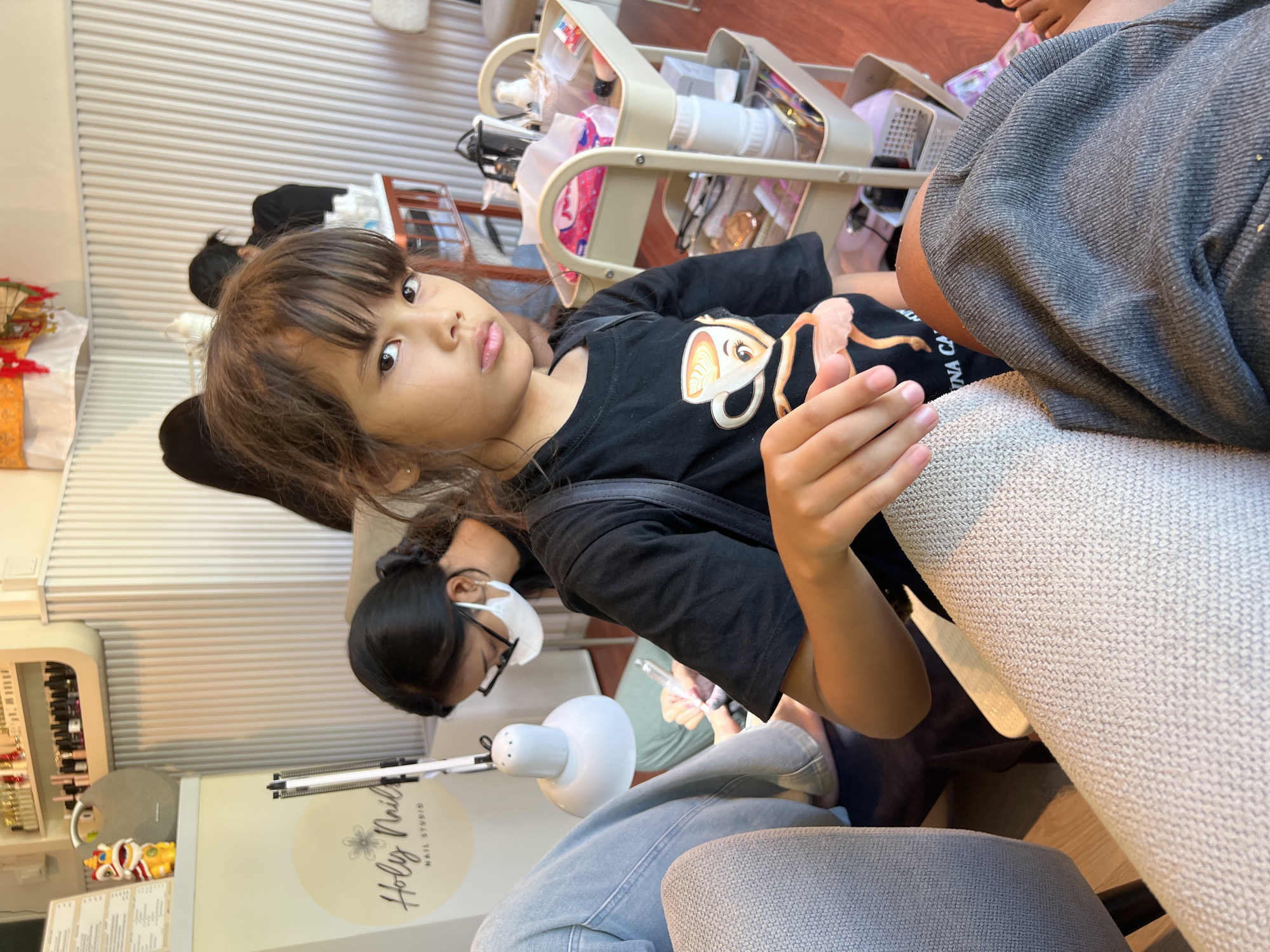A young girl with dark hair and bangs, wearing a black t-shirt with a coffee cup graphic, sits in the foreground of the image. She is looking directly at the camera with a neutral expression. Her right hand rests on a textured light-colored fabric, likely a chair or cushion. In the background, a woman wearing a black t-shirt and glasses is seen working on someone's feet. She is wearing a white mask and holding a tool, suggesting a nail salon environment. A nail lamp is positioned over the feet. Other individuals and a multi-tiered trolley with various beauty products are also visible in the background, indicating a busy salon setting. The overall environment appears to be a well-lit indoor space, possibly a nail studio in Gianyar, Indonesia, given the "Holy Nail Studio" logo visible on signage.