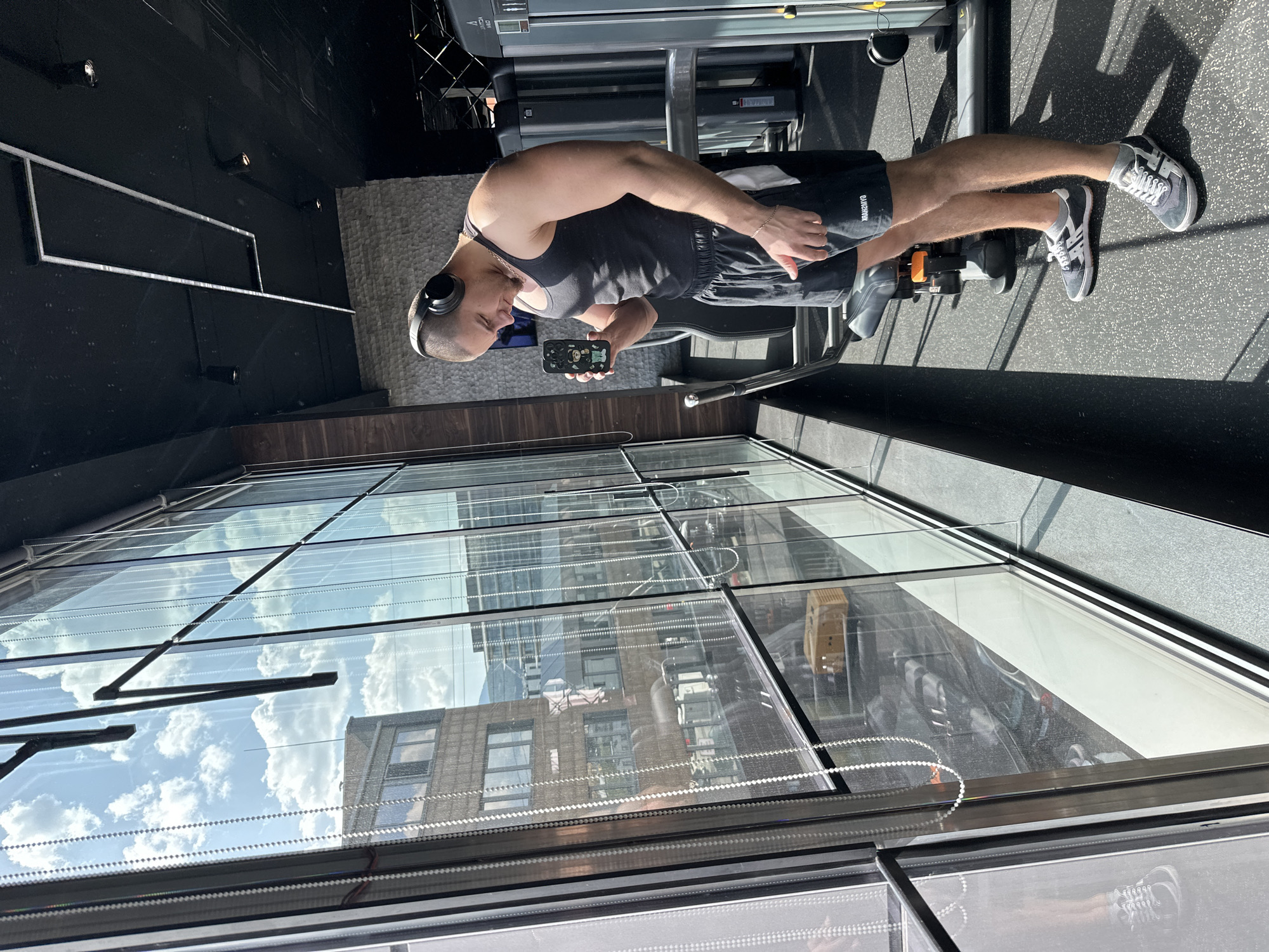 The image depicts a man standing in what appears to be a gym or fitness center in Sofia, Bulgaria. He is seen from the waist up, facing slightly left, looking down at his smartphone. He is wearing a dark grey or black sleeveless tank top, dark shorts with a partially visible logo (possibly "GANNONXX"), and grey and white sneakers with white socks. Black over-ear headphones rest on his head, and a thin bracelet is on his left wrist.
The man holds a smartphone with a distinctive dark case featuring several colorful, cartoon-like characters, including a green frog, a white ghost, and others. He appears to be taking a break from exercising, focused on his phone screen. Behind him, gym equipment like a leg press or similar machine is visible, along with a rower in the foreground near his feet, bearing the text "GYY."
The setting is bright, indicating daytime. Large, clear windows occupy the background, reflecting a bright blue sky with white clouds and several urban buildings. One prominent reflection shows a multi-story building made of brown brick with numerous windows. The interior features a dark floor, a textured grey wall, and a dark ceiling with recessed lighting. Wooden paneling runs along the base of the windows. The sunlight casts distinct shadows on the gym floor.