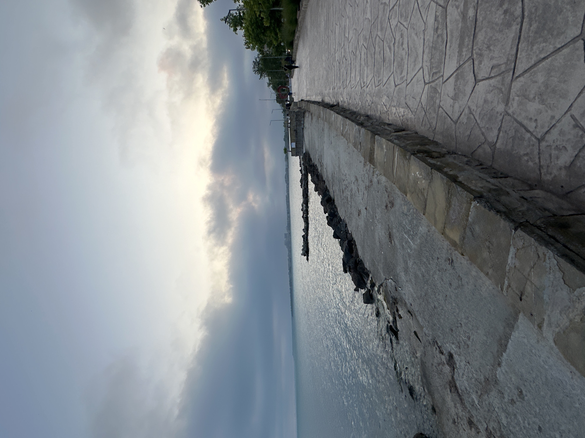 The image depicts a waterside promenade or walkway under a heavily overcast sky, likely at dusk or dawn. The location is in the city of Unknown, Unknown.
On the left, a large body of water, possibly a lake or sea, extends to the horizon, reflecting the grey sky. A rocky breakwater extends into the water from the left foreground, transitioning into a rough, grey concrete platform that lines the shore. In the far distance across the water, a faint urban skyline is visible under a haze.
A low retaining wall, constructed from irregularly shaped, stacked stone blocks, separates the concrete platform from a wide, paved walkway on the right. The walkway features a distinct grey, irregular polygonal stone pattern. Multiple streetlights line the right edge of the walkway, beyond which are green trees and bushes.
Several individuals are present on the walkway in the mid-ground. One person in dark clothing walks away from the viewer. Further down the path, a prominent red-orange circular object, possibly an art installation or sign, is visible. The sky transitions from dark grey clouds overhead to a brighter, paler grey-white band at the horizon, indicating a low light source obscured by clouds. A small, dark, bird-like silhouette is visible high in the lighter sky on the right. The overall lighting is dim and diffused.