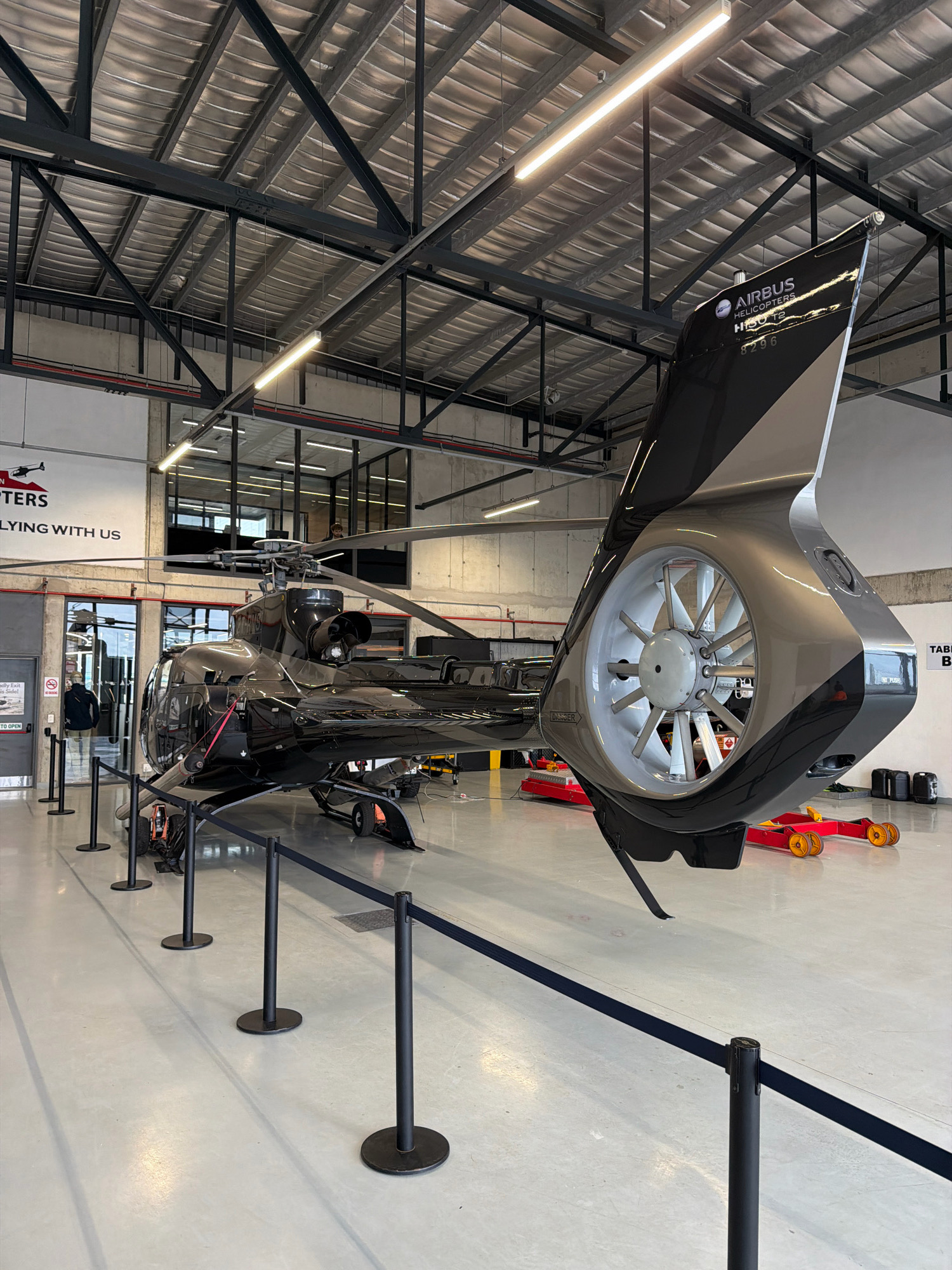 The image shows the rear section of a black helicopter with a silver and black fin. The helicopter is indoors, likely in a hangar, with a polished concrete floor and a high ceiling with exposed metal beams and fluorescent lighting. The helicopter is on display and cordoned off with black stanchions and a dark blue velvet rope. In the background, a large sign with "AIRBUS HELICOPTERS" and "FLYING WITH US" is visible. To the left, a doorway shows a person walking inside. To the right, a "TABLE B" sign and some black cases are present. The overall scene suggests a viewing area or exhibit within an airport or aviation facility. This location is likely the Cape Town International Airport in Cape Town, South Africa, given the presence of Airbus Helicopters operations in the region.