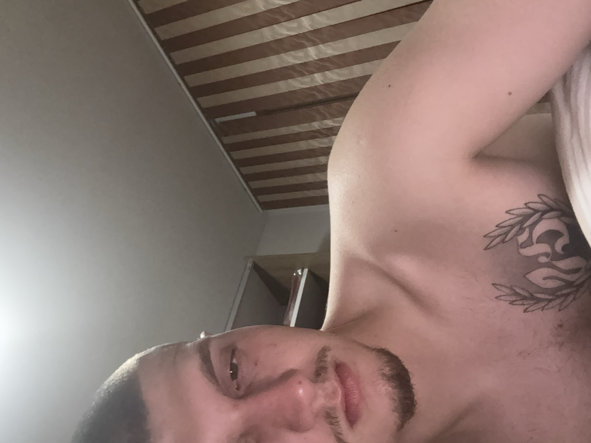 The image displays a male individual, observed from the mid-chest upwards, in an indoor setting identified as a location in Blagoevgrad, Bulgaria. The subject, a male with a light complexion, possesses short, dark hair, a mustache, and a goatee. His left eye is partially closed, and his expression is neutral. A prominent tattoo, appearing to be a laurel wreath encompassing the stylized number "25" or similar emblem, is present on his upper left chest. He is shirtless. The background to the left and top features a plain, light-colored ceiling. To the subject's right, an open shelving unit with two visible shelves is present. To his far right and behind his left shoulder, a wall is covered with vertical, alternating light and dark brown striped blinds or wallpaper, topped by a white trim line where it meets the ceiling. The immediate foreground shows a white, possibly striped, textile, likely bedding. The scene is illuminated by ambient indoor lighting.