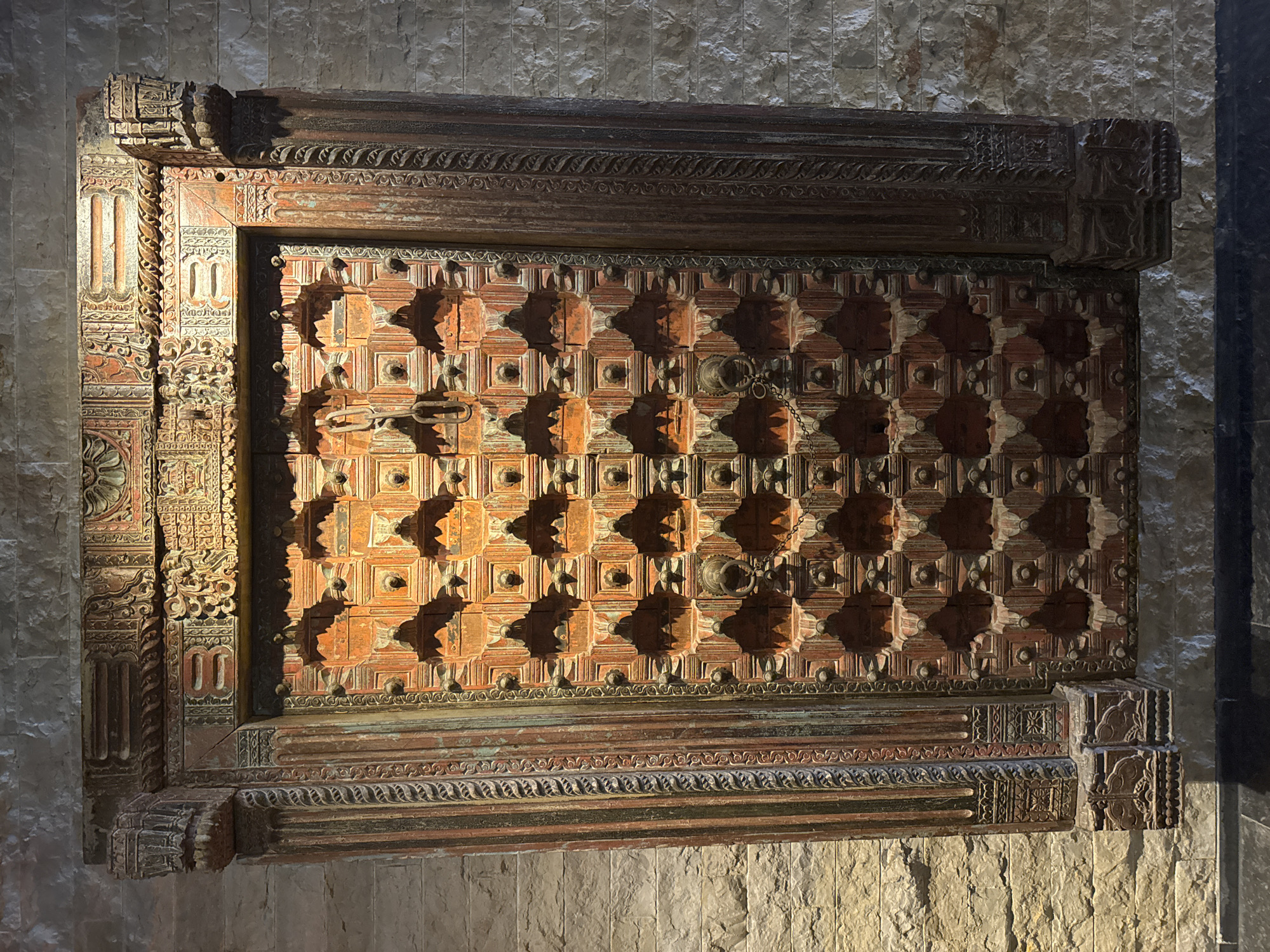 A close-up shot displays an intricately carved, rectangular wooden door or panel, attached to a stone wall. The panel features a repeated square pattern, each square embossed with a central circular knob-like detail, creating a pattern of shadows. The door is framed by a highly ornamented border. The wall, textured in an irregular, stone-like pattern, suggests an interior setting. Light source on the left side of the door creates shadow, and the location is not found.