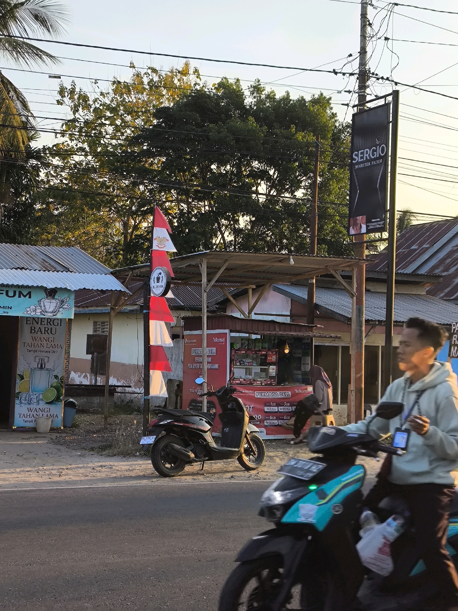 In Tenilo, Indonesia, a roadside scene unfolds under the warm glow of the setting sun. A man, wearing a light blue hoodie and dark pants, rides a turquoise scooter with a black front. He holds a mobile device in his right hand, and a plastic bag hangs from the front of his scooter. Another black scooter is parked on the side of the road.
Adjacent to the road is a small building with a corrugated metal roof, housing a shop. A red and white Indonesian flag flutters from a pole in front of the shop. Signs advertising "SERGIO 20 KRETEK FILTER" and a perfume brand with images of bottles and citrus fruits are visible on poles and the shop's facade. A person wearing a hijab is seen inside the shop. The environment is characterized by power lines crisscrossing overhead and trees with green foliage in the background. The road surface is asphalt, with some debris and vegetation along the roadside.