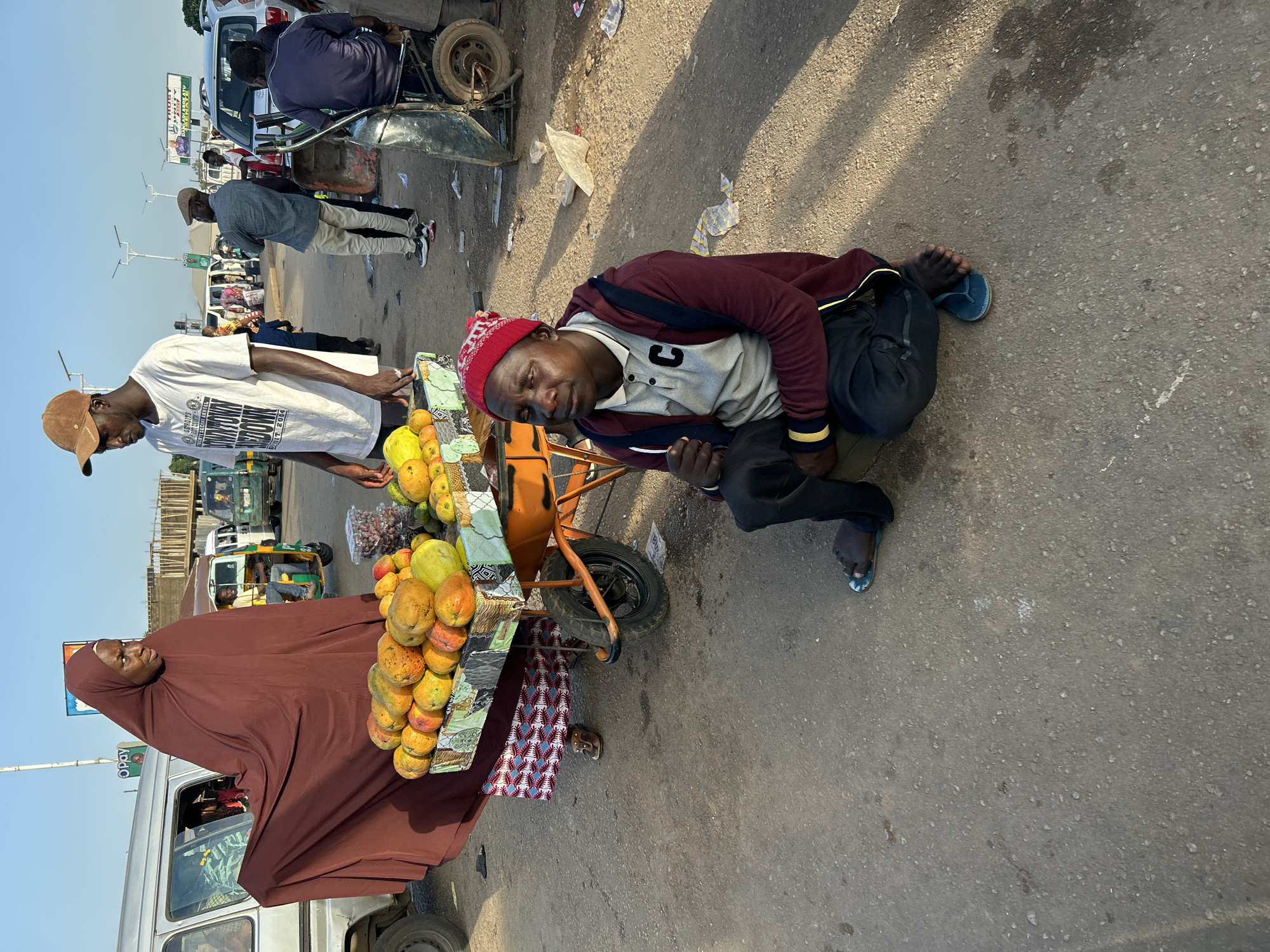 The scene is an outdoor market in Jos, Nigeria. A woman wearing a maroon hijab stands next to a fruit cart, which displays mangoes and grapes. A man in a white t-shirt and brown cap is near the cart. Another man is kneeling on the ground near the cart and wearing a maroon jacket, gray shirt, and red hat. In the background, there are vehicles, including cars, vans, and motorcycles, and several other people milling about. There is also a sign with various words, including “TROY” and “SUPREME”. The ground is paved with asphalt and has some debris.