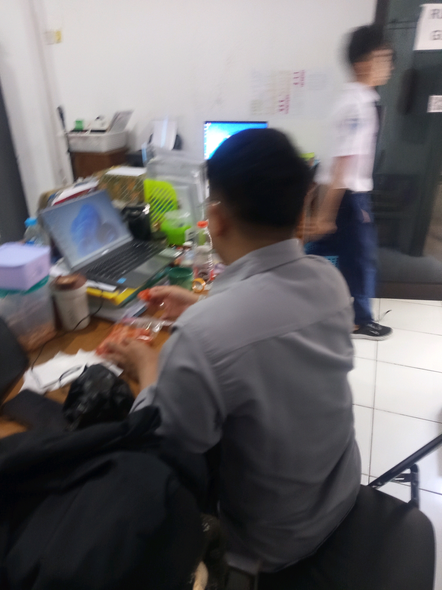 This is a slightly blurred, low-resolution image taken indoors, likely in an office or classroom setting. In the foreground, a person wearing a gray collared shirt is seated, with their back to the camera, appearing to be holding a plastic bag containing orange items. Their hands are visible. In front of them is a wooden desk with a laptop, some containers, and a bottle of liquid. In the background, another person, wearing a white shirt and dark pants, is walking away from the camera. The environment appears to be a brightly lit room with white walls. There are papers and objects on the walls and a shelf unit with items. The floor is tiled. The image context suggests a typical day in a professional or educational institution in Malang, Indonesia.