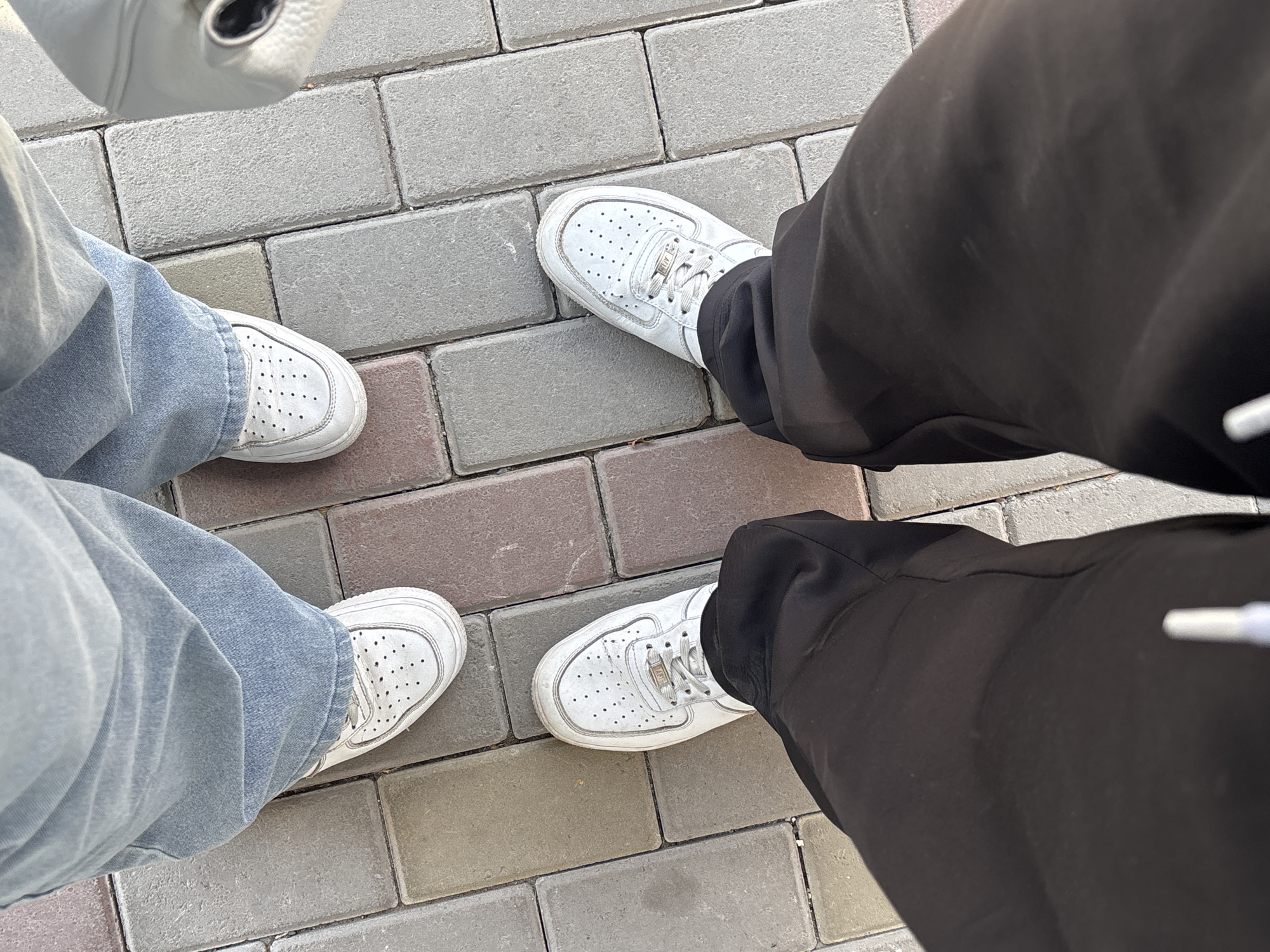 The image shows a view from above of two pairs of legs standing on a paved surface. The paving consists of rectangular blocks. On the left, the person is wearing faded blue jeans and white sneakers. On the right, the person is wearing black pants and white sneakers. The location is Yerevan, Armenia.