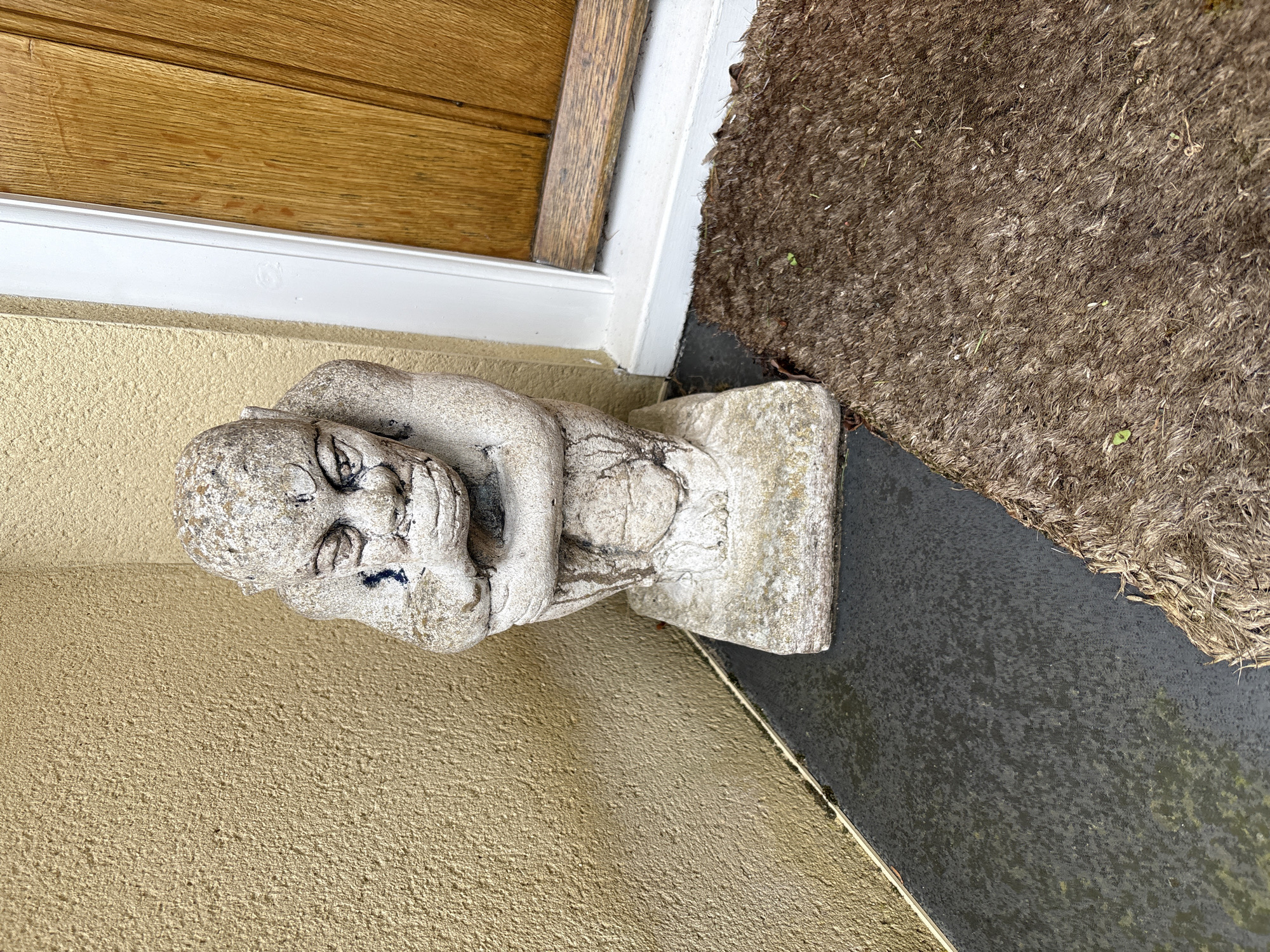 A stone gargoyle sits next to a doorway in Longden, United Kingdom. The gargoyle, weathered and showing signs of age with dark stains, is positioned on a textured, tan-colored wall adjacent to a white door frame and a light brown wooden door. A brown, rough-textured doormat is also visible. The photograph is taken from a close, elevated angle, revealing details of the surfaces and textures of the objects.