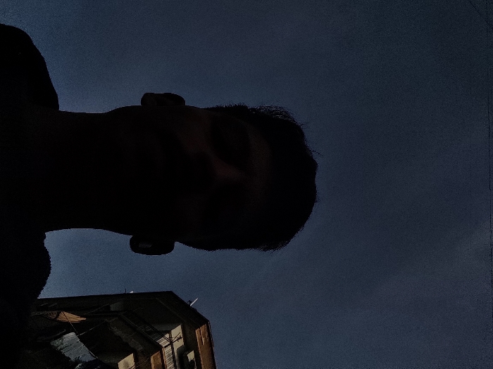 The image presents a low-angle shot, featuring a silhouetted individual in the foreground against a dark blue sky, located in Al-Qanawat Municipality, Syria. The subject, a person with discernible head and shoulders, is positioned centrally, facing slightly upwards, their features obscured by strong backlighting. To the right, a multi-story residential building with light-colored facades and dark-framed windows is partially visible. Laundry is hanging on a line from a lower balcony of this building. Several small, white satellite dishes are affixed to the upper sections of the structure. The sky is clear, with a deep blue hue indicative of twilight conditions.