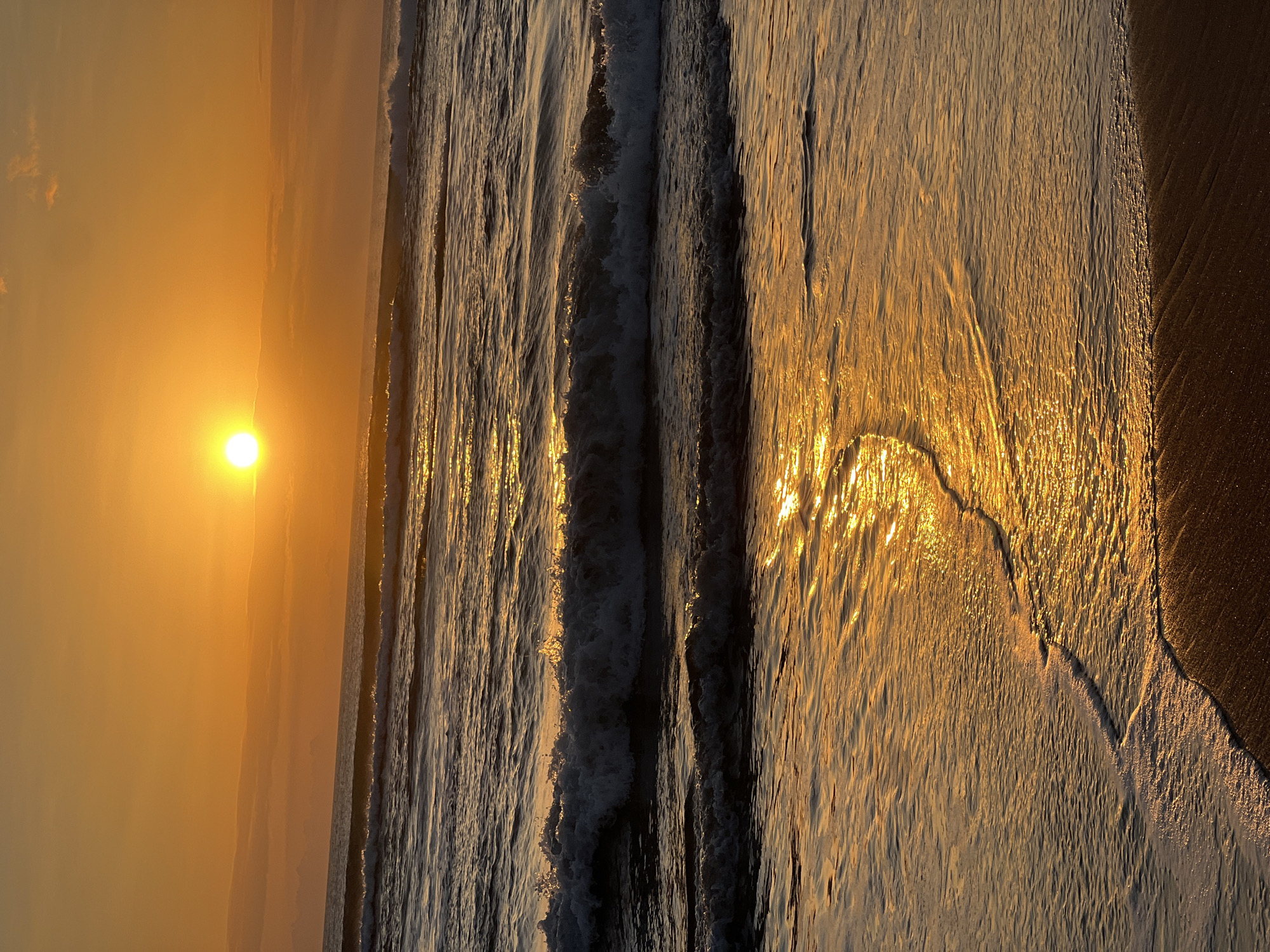 A low-angle, vertical shot captures the ocean at Kuta Beach, Indonesia, during sunset. The sun, a bright, glowing orb, sits low in the sky, casting a warm, golden-orange hue across the scene. Wispy clouds are visible in the distance. Gentle waves roll towards the shore, with the crests of some waves illuminated by the setting sun, creating a shimmering effect on the water's surface. The wet sand at the edge of the beach reflects the golden light, with a thin line of foam from a receding wave gently lapping at the shore. No people or man-made objects are visible in the frame.