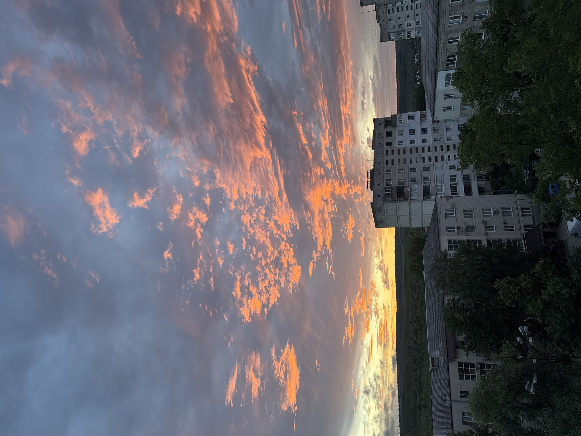 The image depicts an urban landscape in Strășeni, Moldova, dominated by a dramatic sunset. The sky is filled with vibrant orange and pink clouds, interspersed with darker gray clouds, creating a striking contrast. Below the sky, a row of multi-story apartment buildings, typical of Eastern European urban architecture, are visible. These buildings are constructed from concrete and brick, with numerous windows and balconies. Trees and greenery are scattered between and around the buildings, some appearing dense and lush. In the distance, rolling hills covered in vegetation form the horizon line. The overall scene conveys a sense of twilight in a residential area, with the natural beauty of the sunset contrasting with the man-made structures. There are no visible people or ongoing actions.