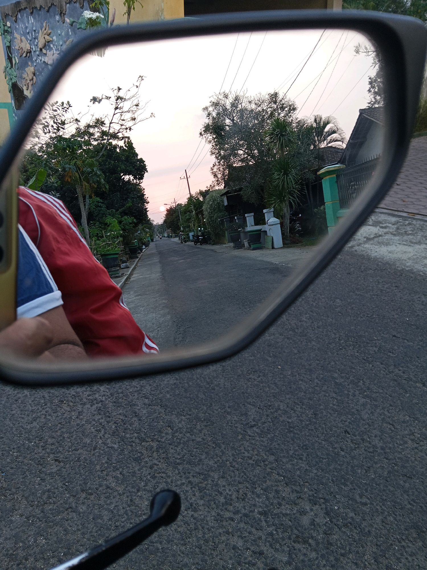 The image is a reflection in a motorcycle's rearview mirror, showing a street scene in Gogodeso, Indonesia. In the foreground of the reflection, a person wearing a red jacket with white stripes and a blue shirt is visible from the chest down. The street appears to be paved with asphalt and lined with trees and houses. Utility poles and power lines are visible overhead. The sky is a soft pink and orange, indicating either sunrise or sunset. There are several motorcycles parked along the side of the road, and a few more are seen in the distance. The houses have tiled roofs and fences, typical of a residential area in Indonesia. The reflection also shows decorative elements on a wall in the upper left corner, which appear to be sculpted figures. The environment suggests a peaceful, rural or semi-urban setting.