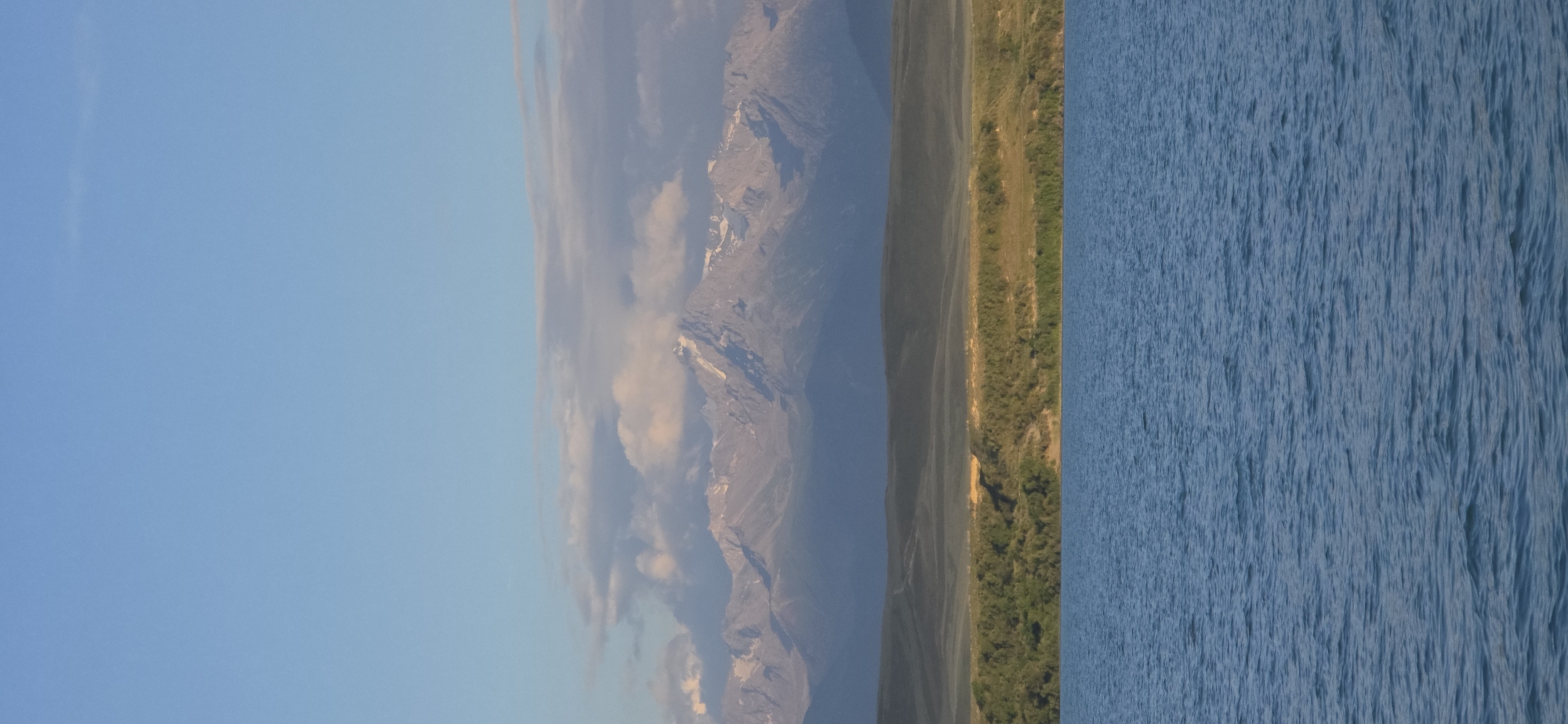A serene landscape captures the vast expanse of a body of water in the foreground, its surface rippling gently under a clear blue sky. Beyond the water, a strip of verdant green foliage lines the shore, giving way to rolling hills that recede into the distance. The horizon is dominated by a majestic mountain range, its snow-capped peaks catching the soft light of the sun, with a scattering of clouds drifting above. The scene evokes a sense of tranquility and natural beauty, typical of the mountainous regions surrounding Lake Issyk-Kul near the city of Cholpon-Ata, in the Issyk-Kul Region of Kyrgyzstan.