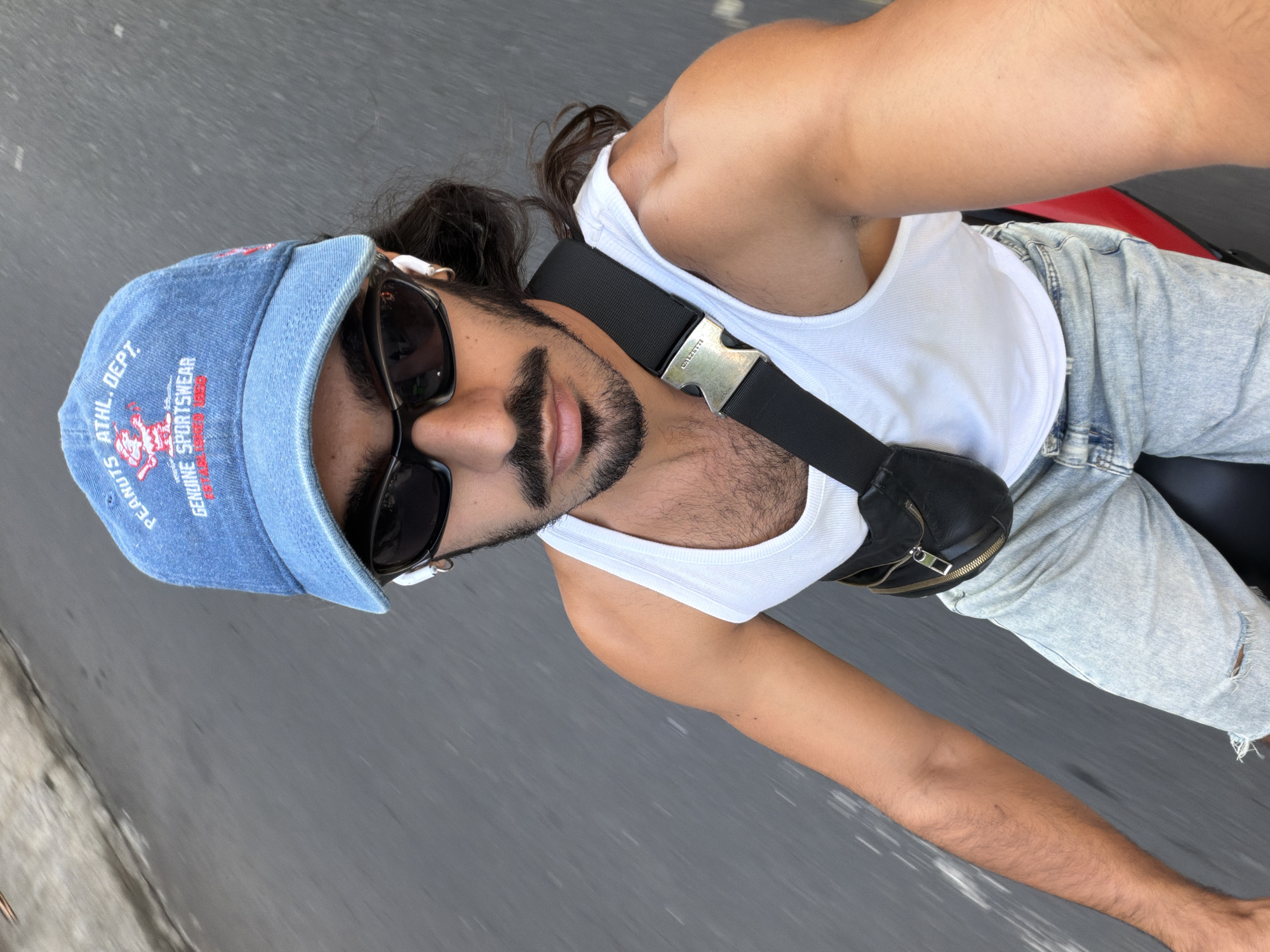 The image captures a first-person perspective of a person riding on a moving vehicle, indicated by the motion blur on the grey road below. The setting is North Kuta, Indonesia. The subject, likely male, is looking towards the camera, wearing dark sunglasses and white wireless earbuds.
They are dressed casually in a white ribbed tank top and light blue, distressed denim jeans. A black fanny pack with a prominent metallic buckle is slung across their chest; the buckle features the engraved brand name "CALZATTI."
On their head, they wear a light blue denim baseball cap. Visible text embroidered on the cap includes "NUTS ATHL. DEPT." and "GENUINE SPORTSWEAR" in red, along with "PEANUTS" partially visible under the brim. The person has dark hair tied back, a neatly trimmed beard, and a mustache. Their right arm, displaying tanned skin and muscular definition, is visible in the foreground. A small red portion of the vehicle they are on can be seen in the upper right corner. The bright lighting suggests it is daytime with clear weather.